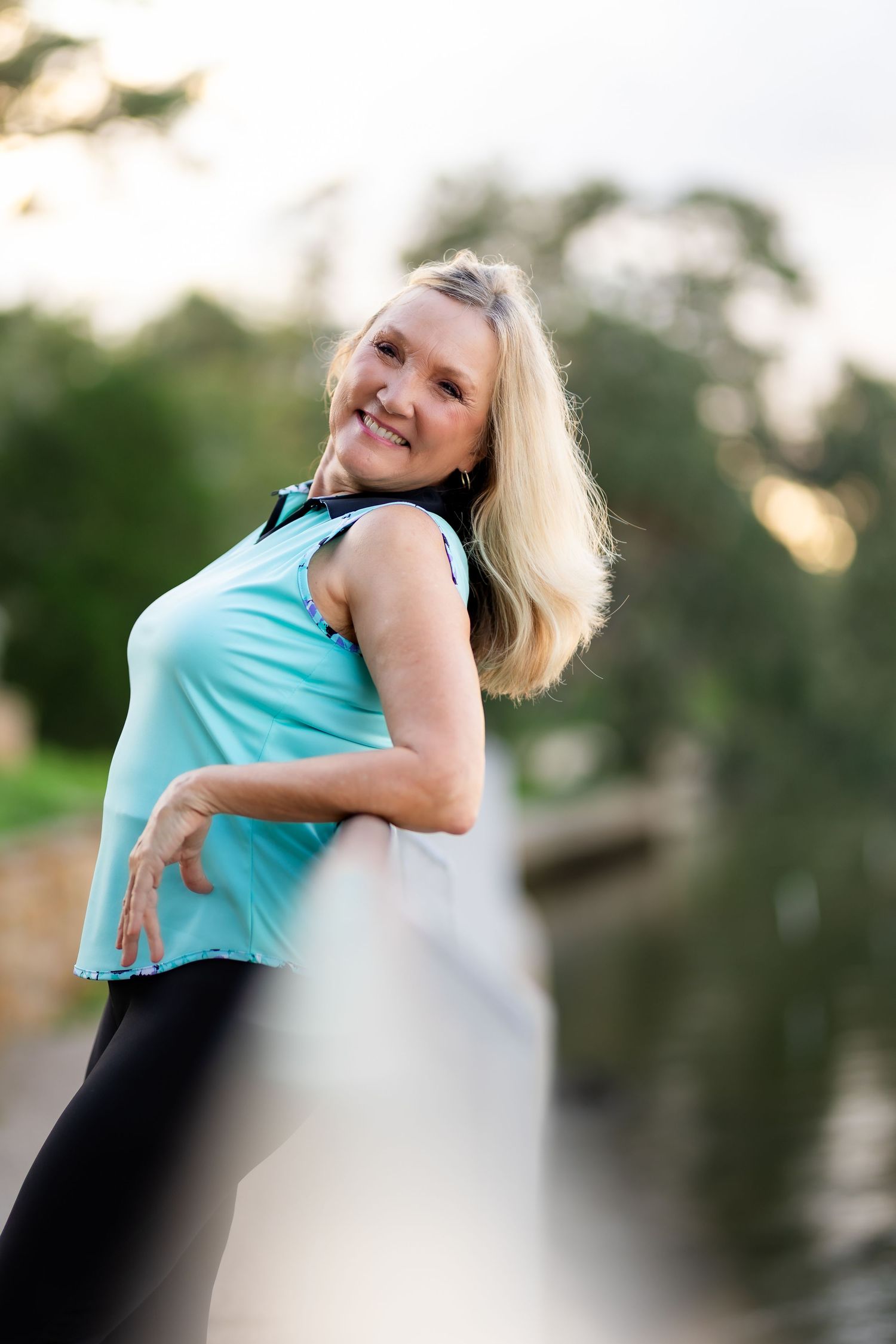 A person in turquoise sleeveless top and dark pants leans back while smiling in an outdoor setting at sunset.