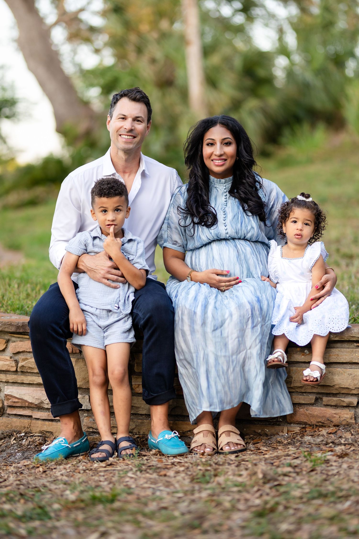 A family wearing coordinated blue and white outfits sits together on a stone wall outdoors during a photo session.