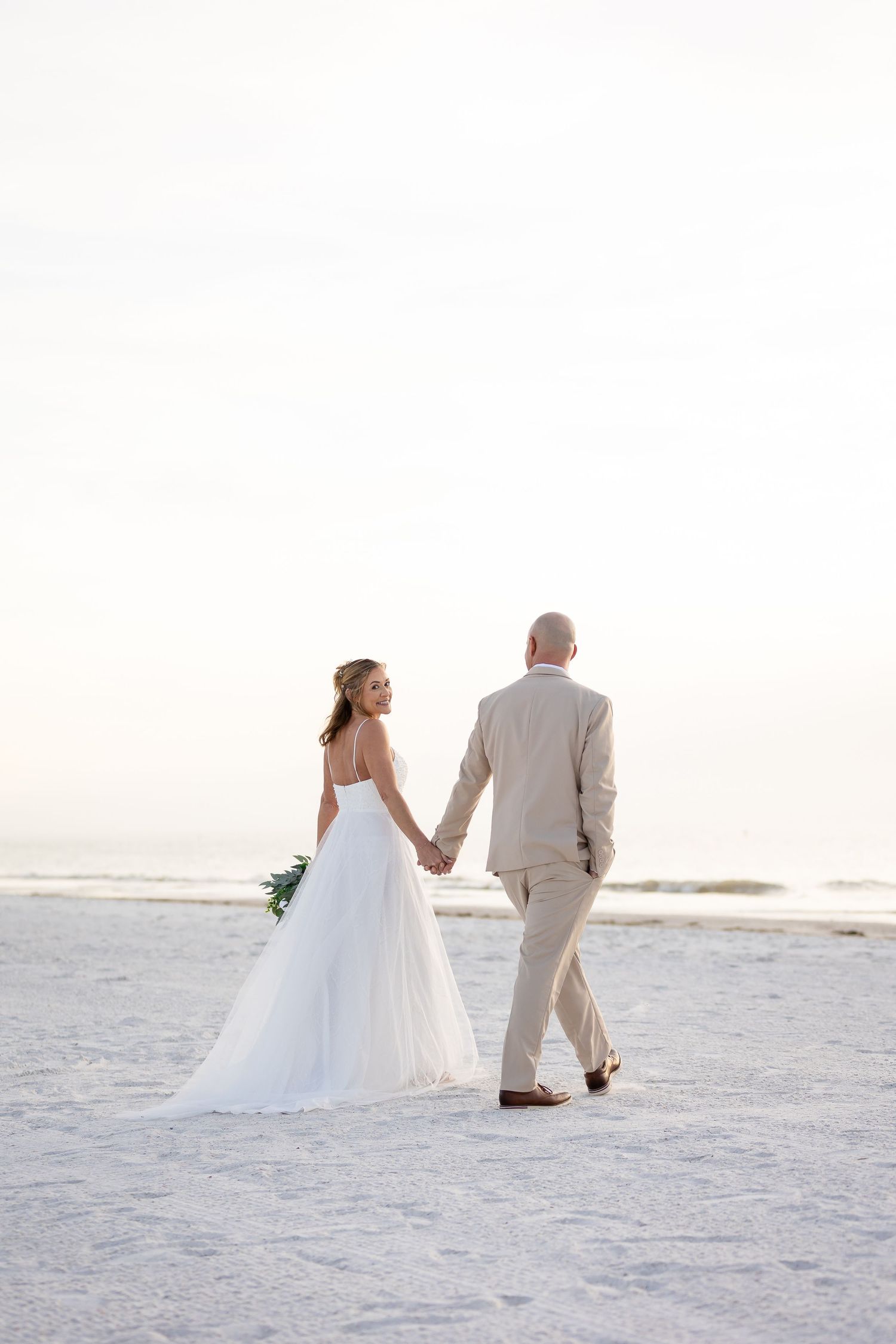 A couple in wedding attire walks hand in hand along a pristine white sandy beach at sunset.