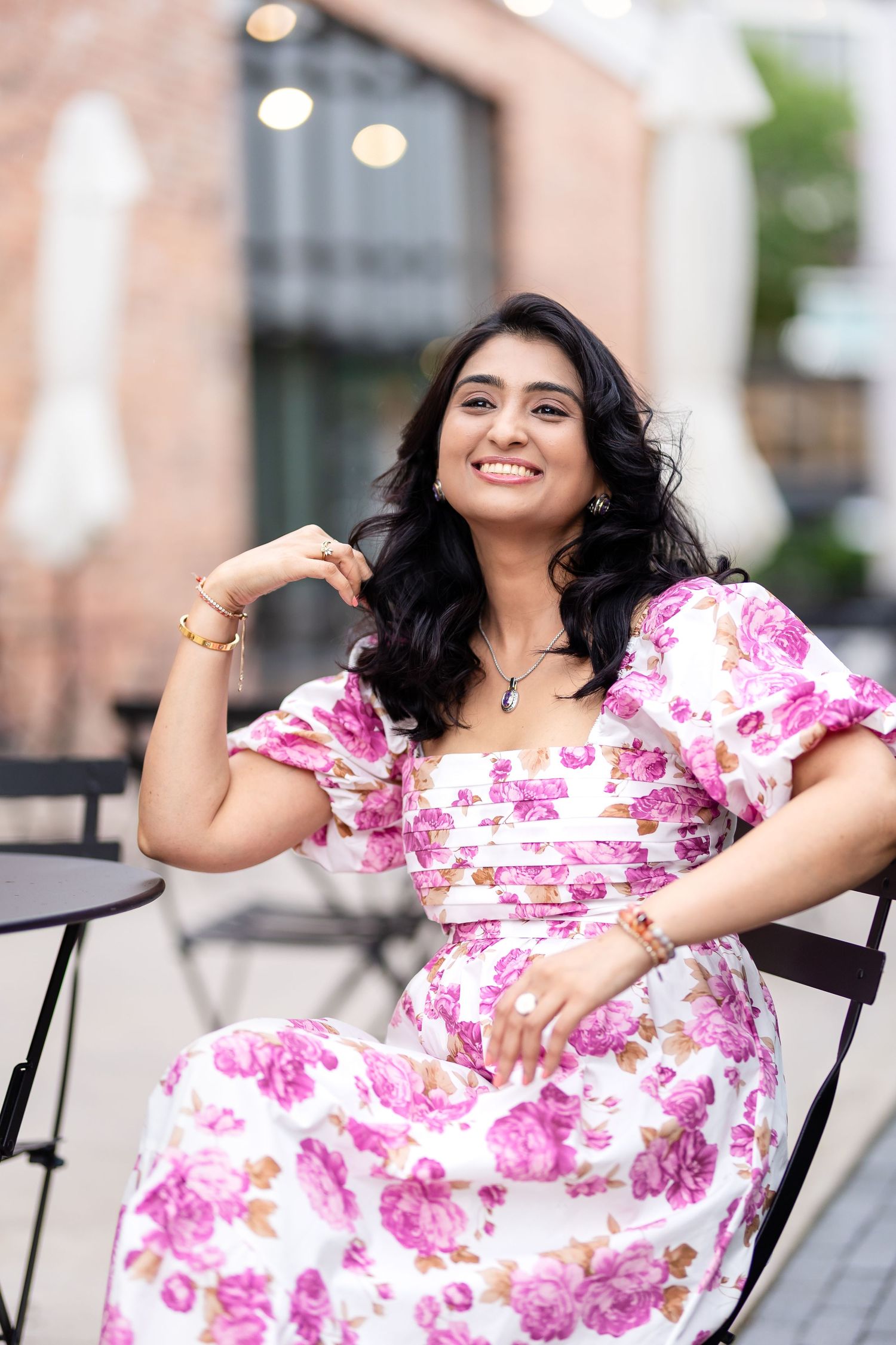 A woman in a pink floral maxi dress smiles while sitting on an outdoor patio chair.