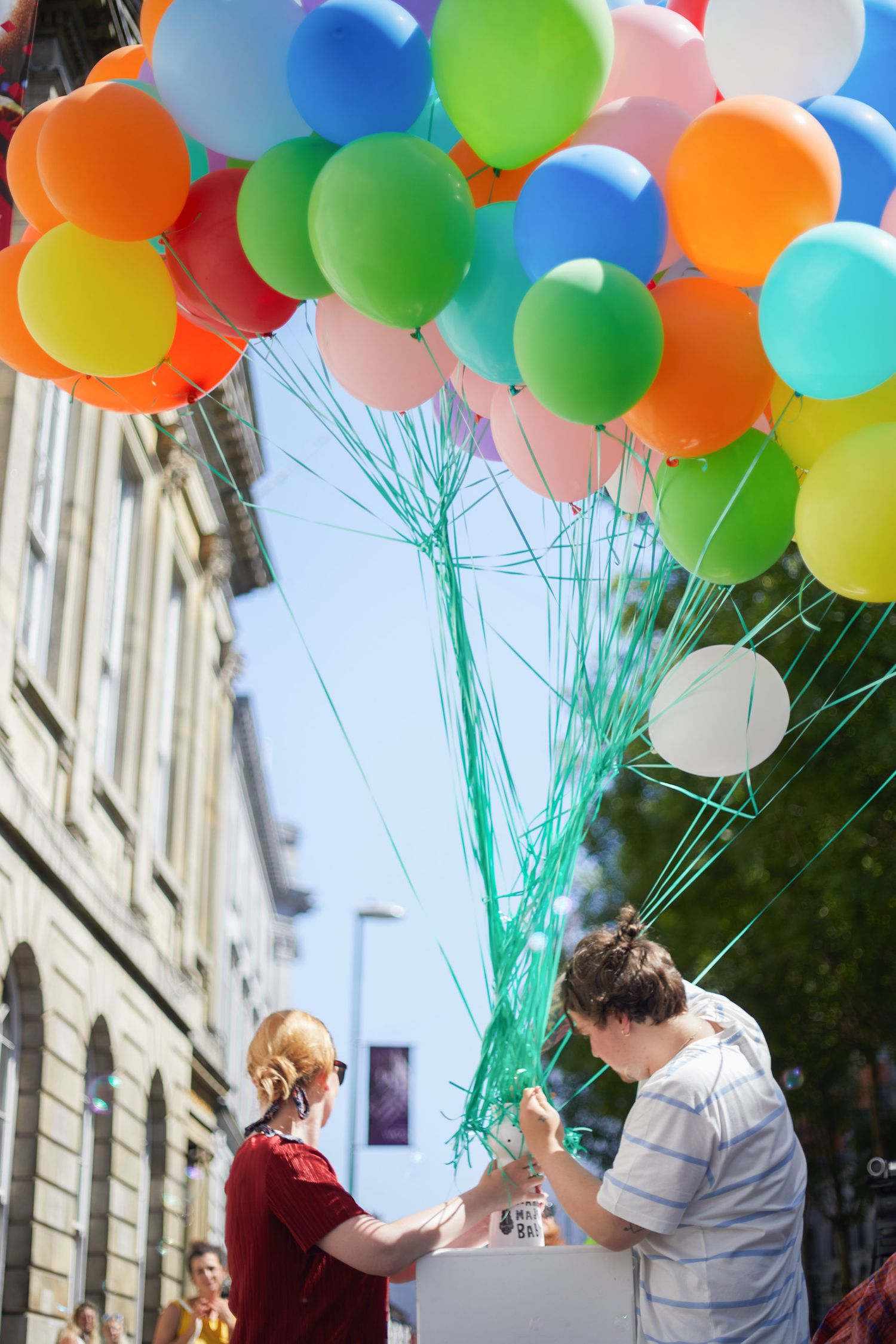 Two people holding colorful helium balloons on a sunny day outside a building.