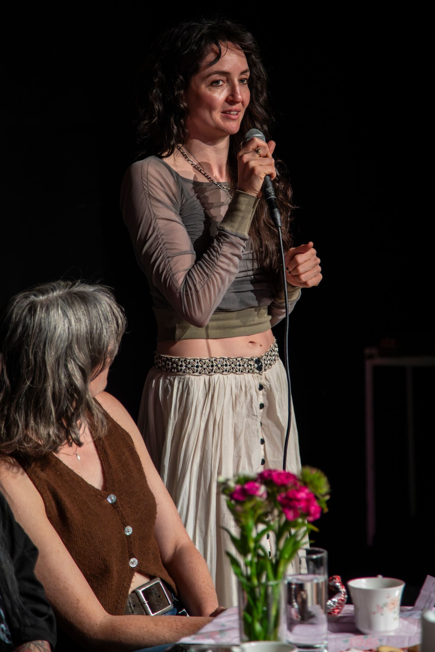 A person in a brown vest and white dress speaks into a microphone on stage with pink flowers visible in the background.