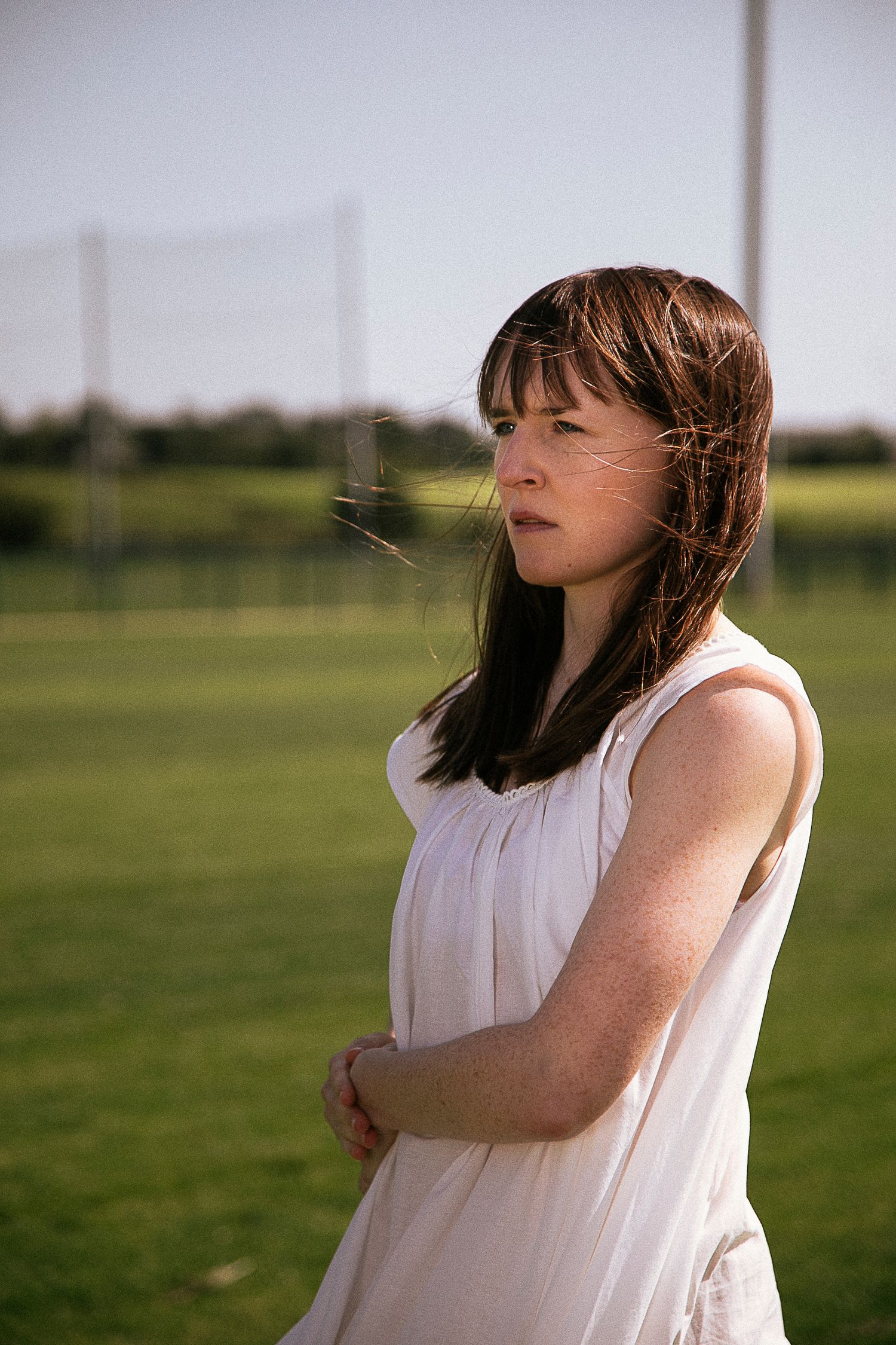 A person in a white sleeveless dress stands in a grassy field with eyes closed as wind blows through their dark hair.