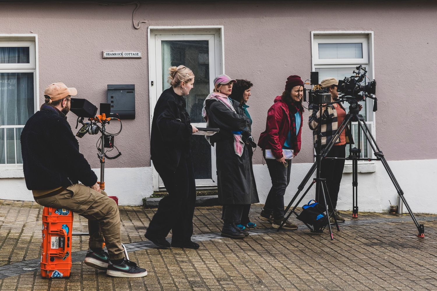 Film crew with cameras and equipment filming outside a pink building on a cloudy day.