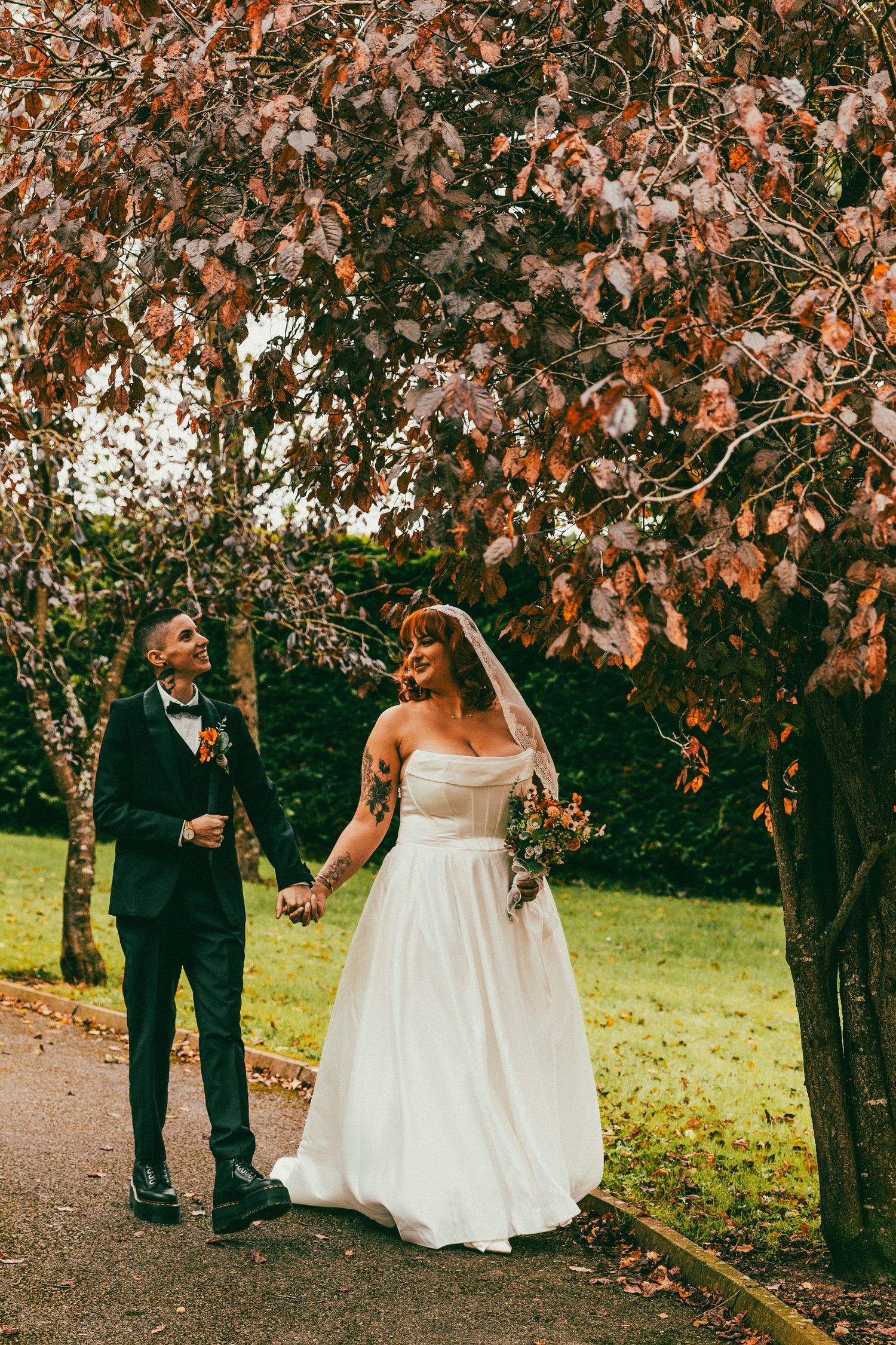 A romantic autumn wedding scene with a couple walking under trees with orange leaves in a park setting.