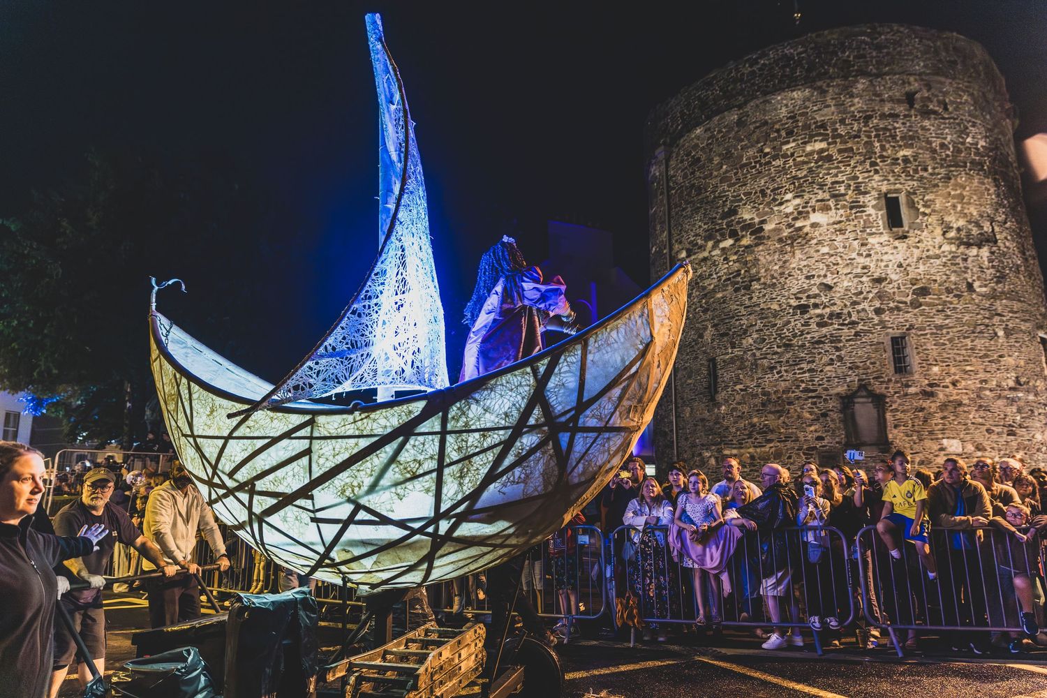 Illuminated castle ruins at night with blue lighting and crowds gathered for an outdoor event.