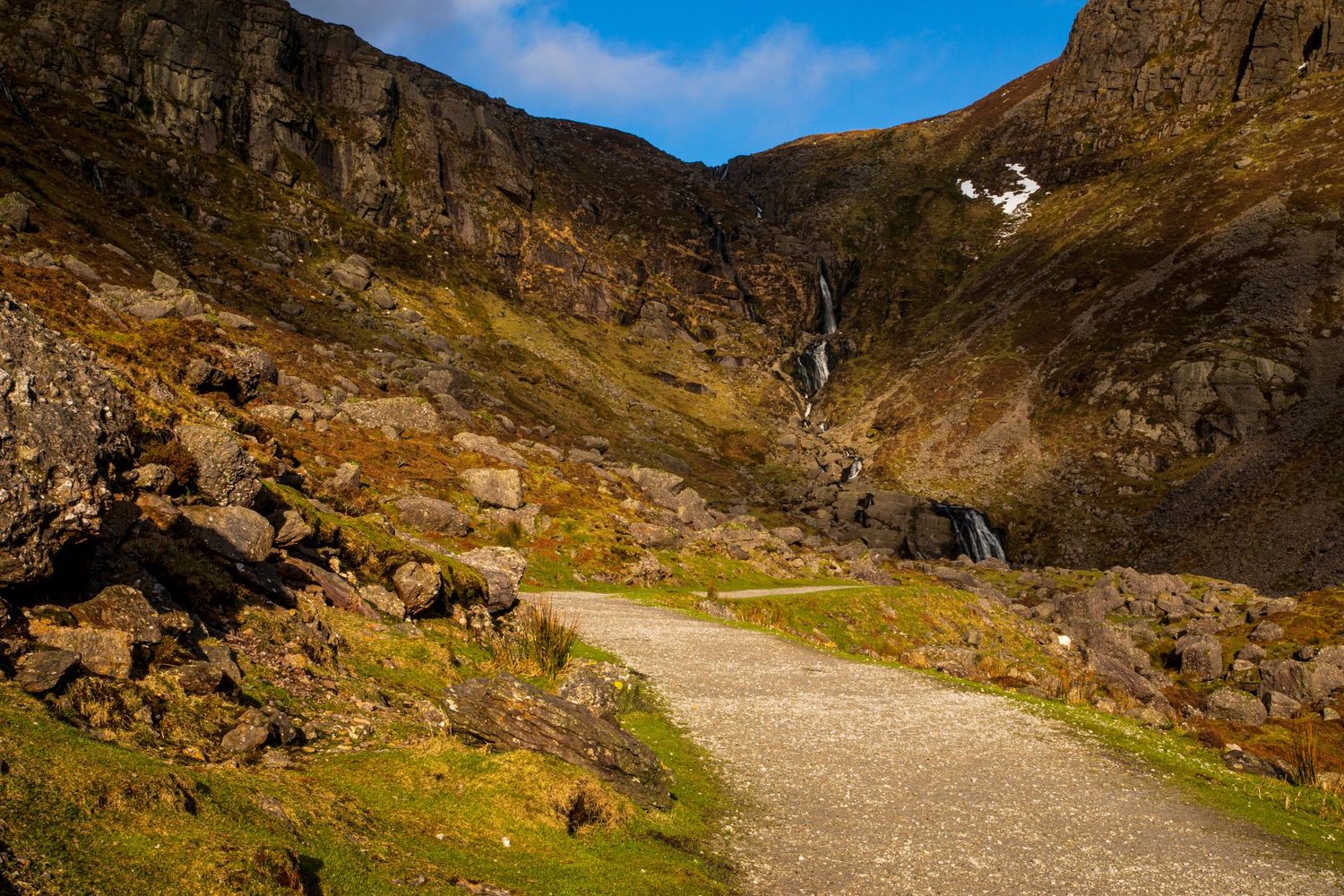 Winding mountain road curves through rugged terrain with steep cliffs and rocky outcrops under blue sky.