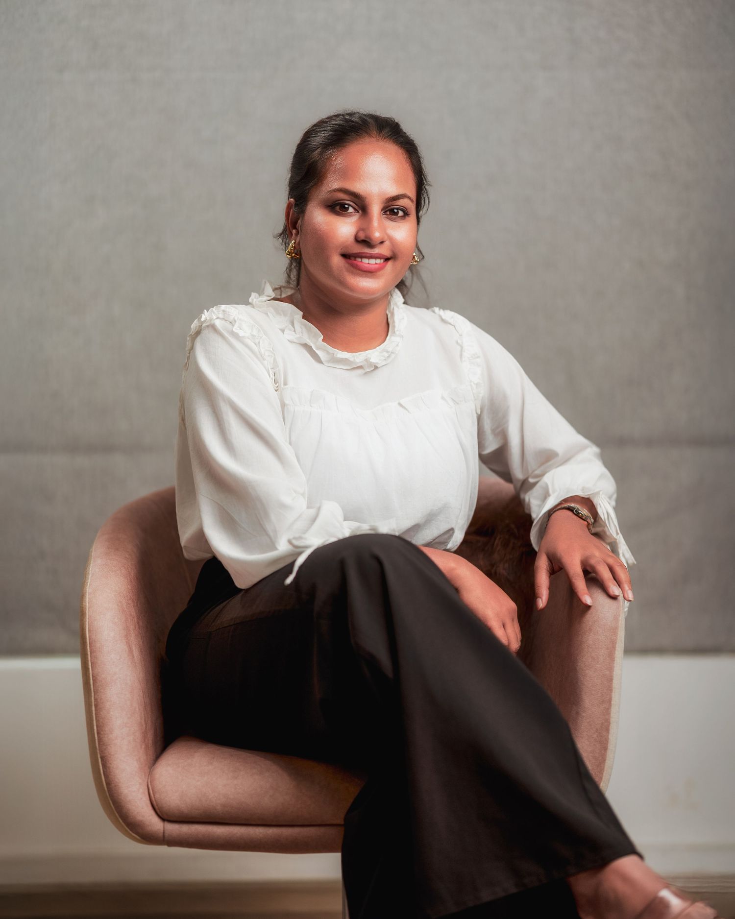 Relaxed professional portrait in white blouse and dark pants on beige chair.