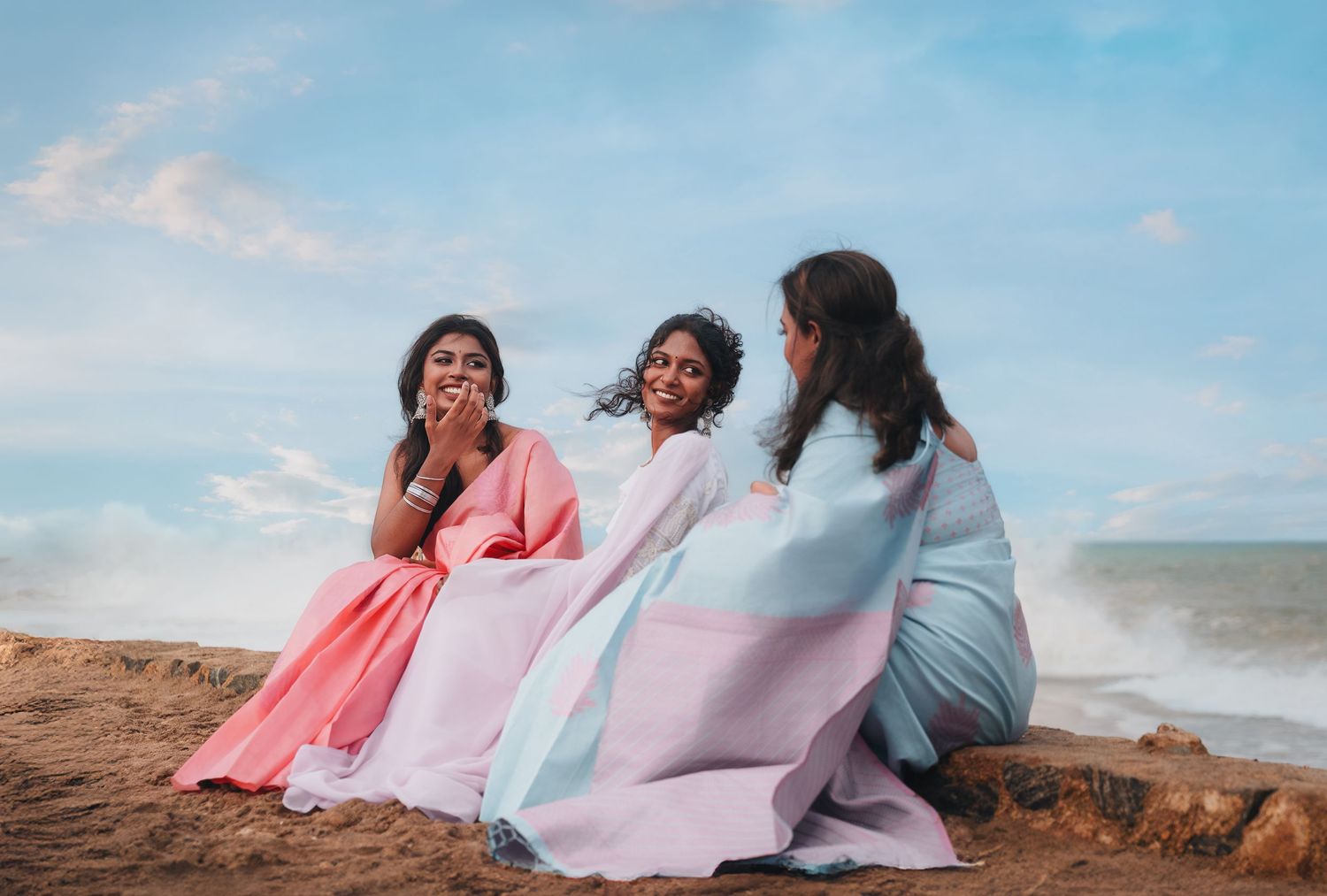 Friends in pastel dresses share laughter while sitting on a beach overlook.