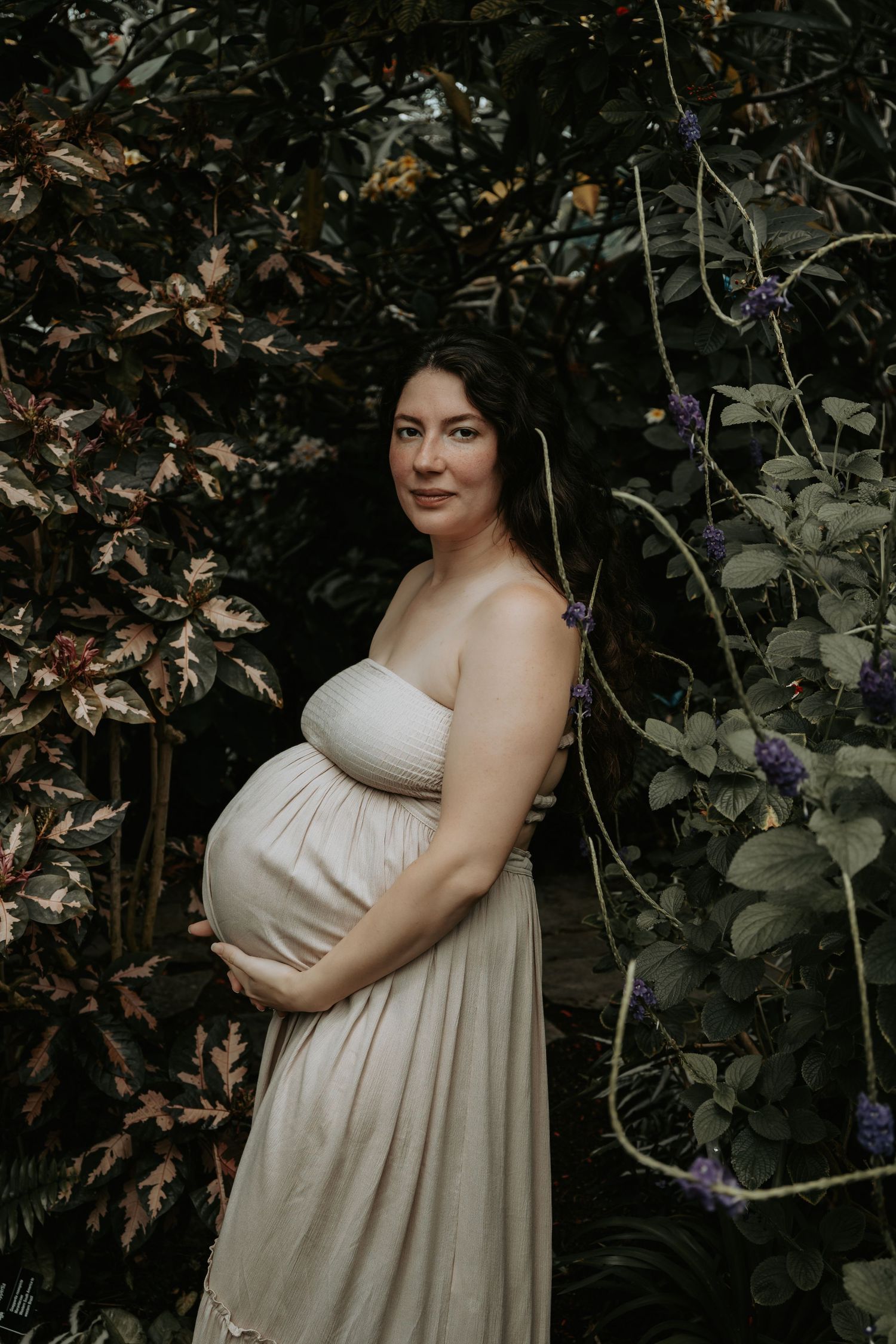 Pregnant woman in cream dress cradling baby bump surrounded by lush green and purple foliage outdoors.