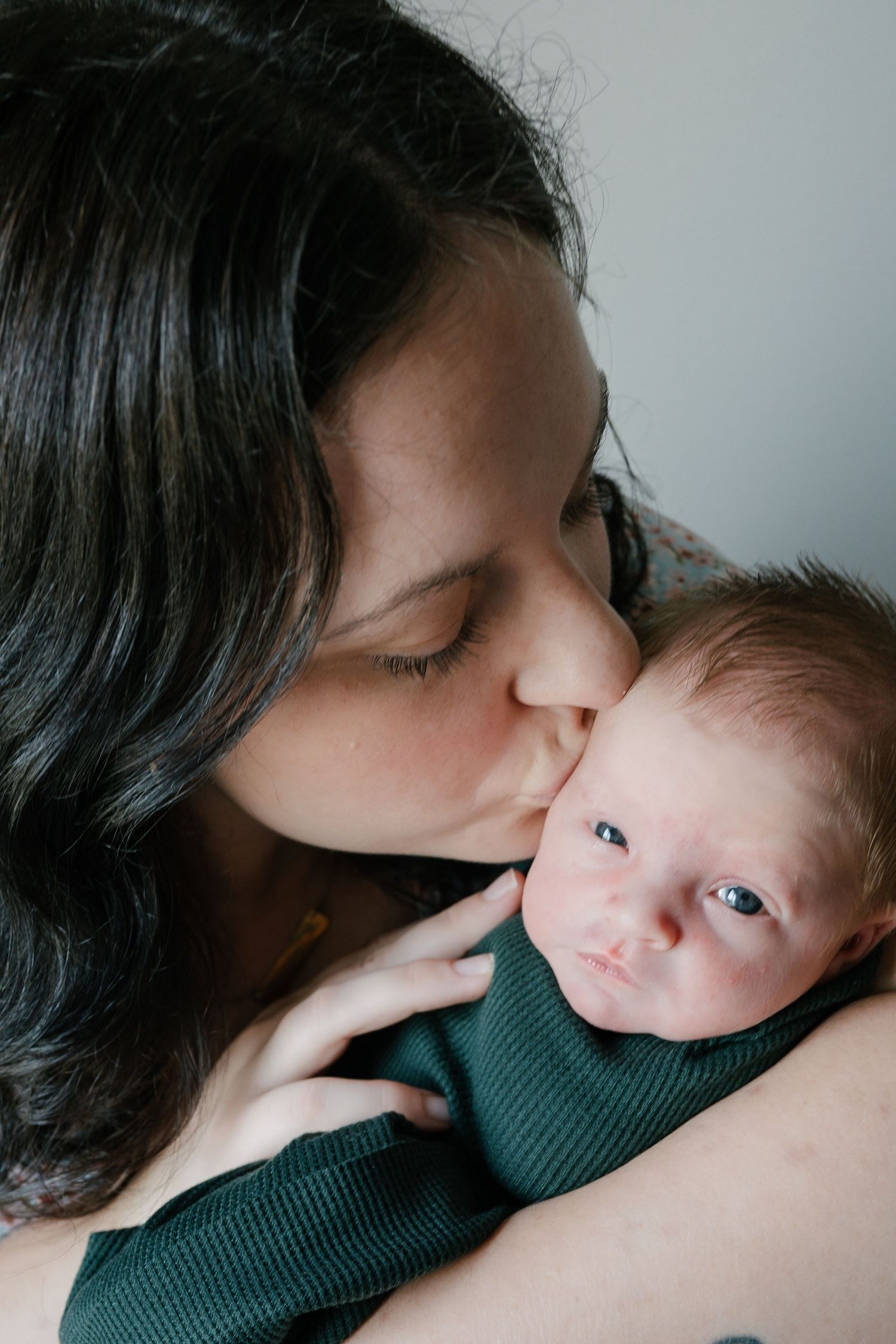 A loving mother with dark hair gently kisses her newborn baby wrapped in a green outfit.