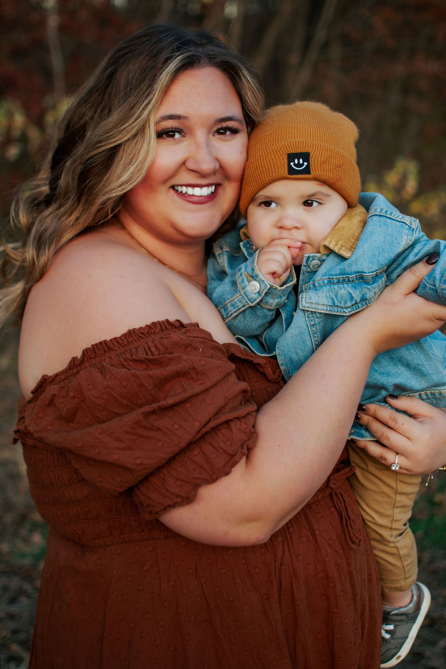 Smiling mom in off-shoulder rust dress holding baby in denim jacket and mustard smiley beanie outdoors.