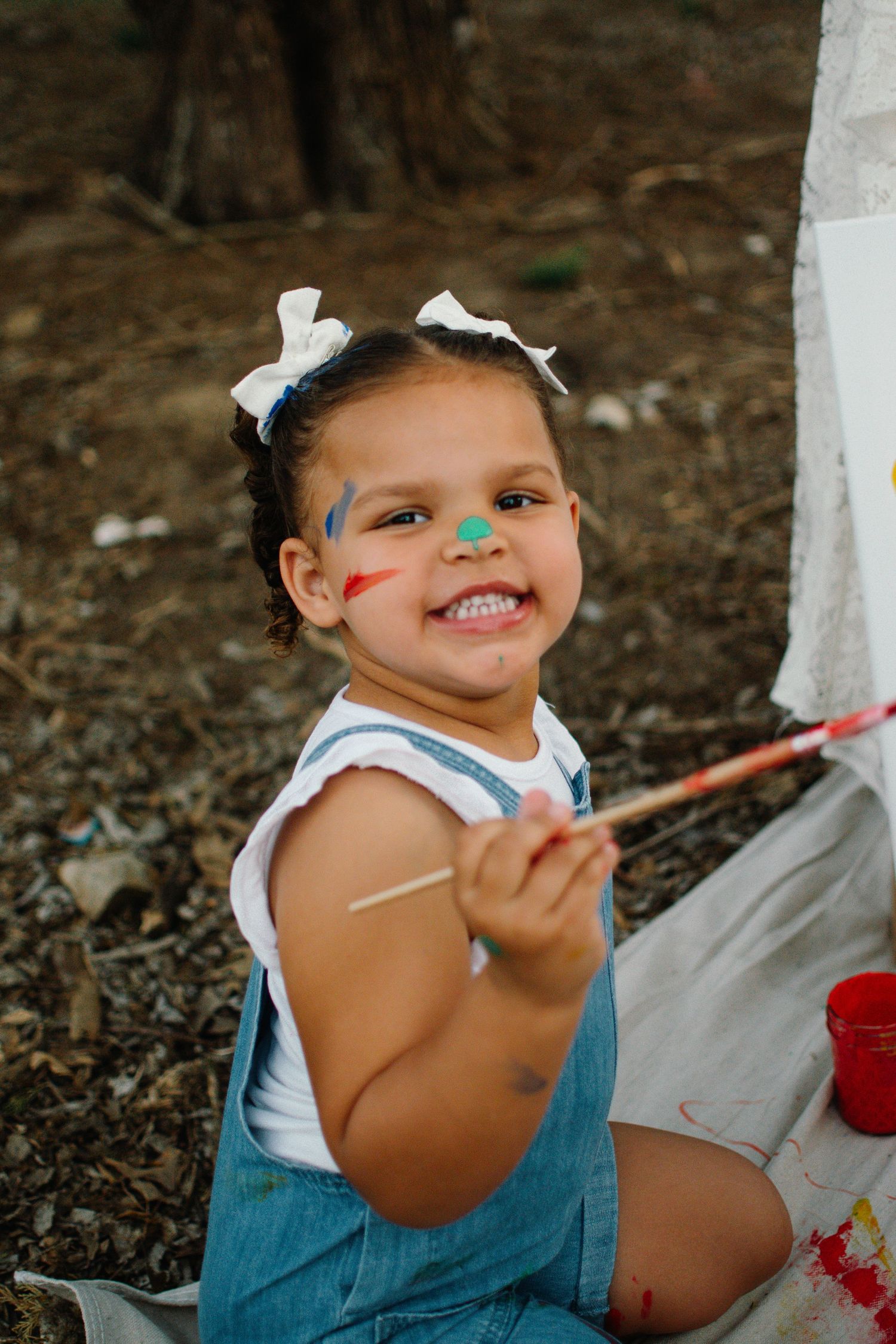 A happy toddler girl with paint on her face holds a paintbrush while doing an outdoor art project.