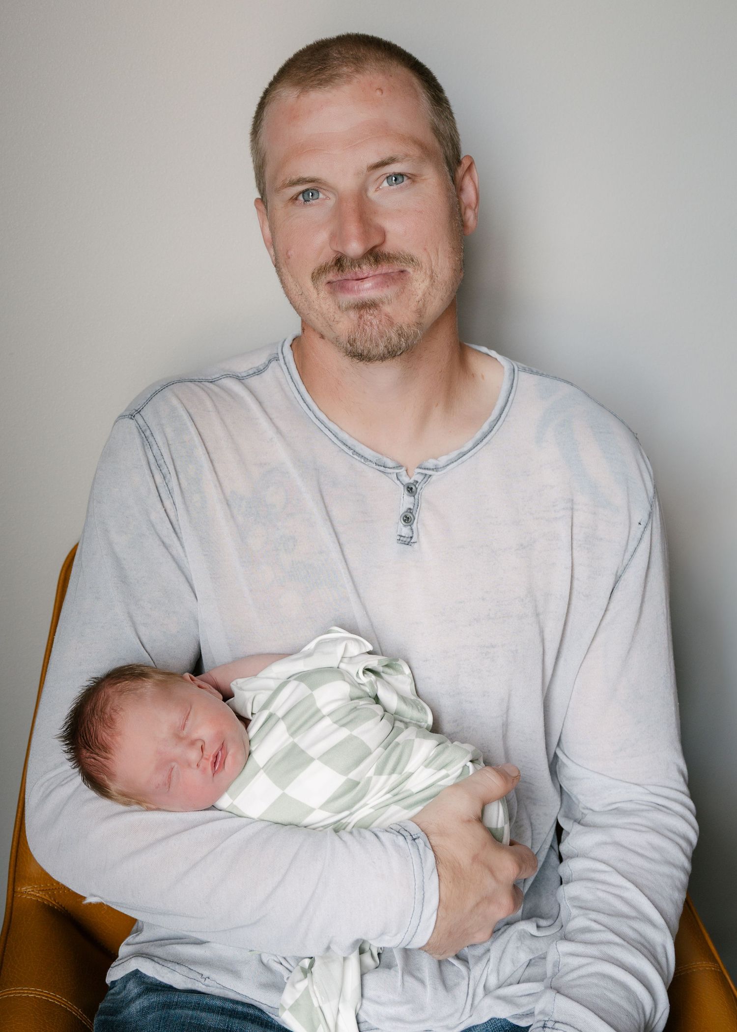 Father holding sleeping newborn baby swaddled in green checkered blanket while sitting in wooden chair.