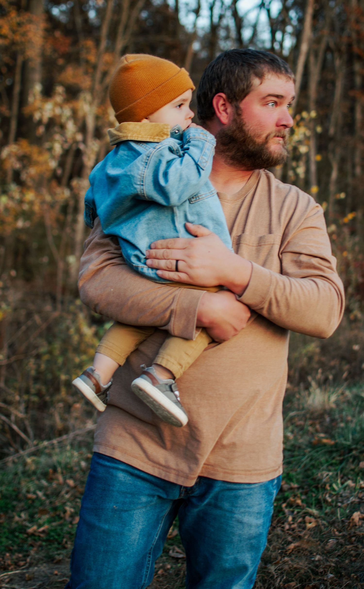 Bearded dad in tan shirt holding toddler in denim jacket and mustard beanie in autumn outdoor setting.