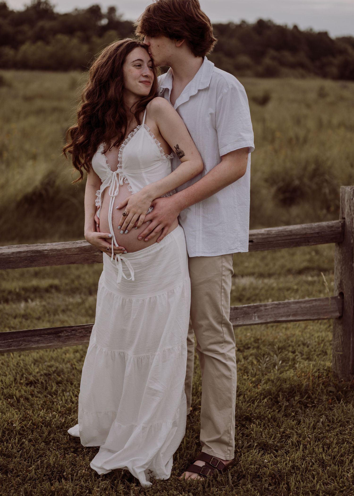 A couple shares a tender maternity photo outdoors, with the man embracing the pregnant woman in a white dress near a wooden fence.