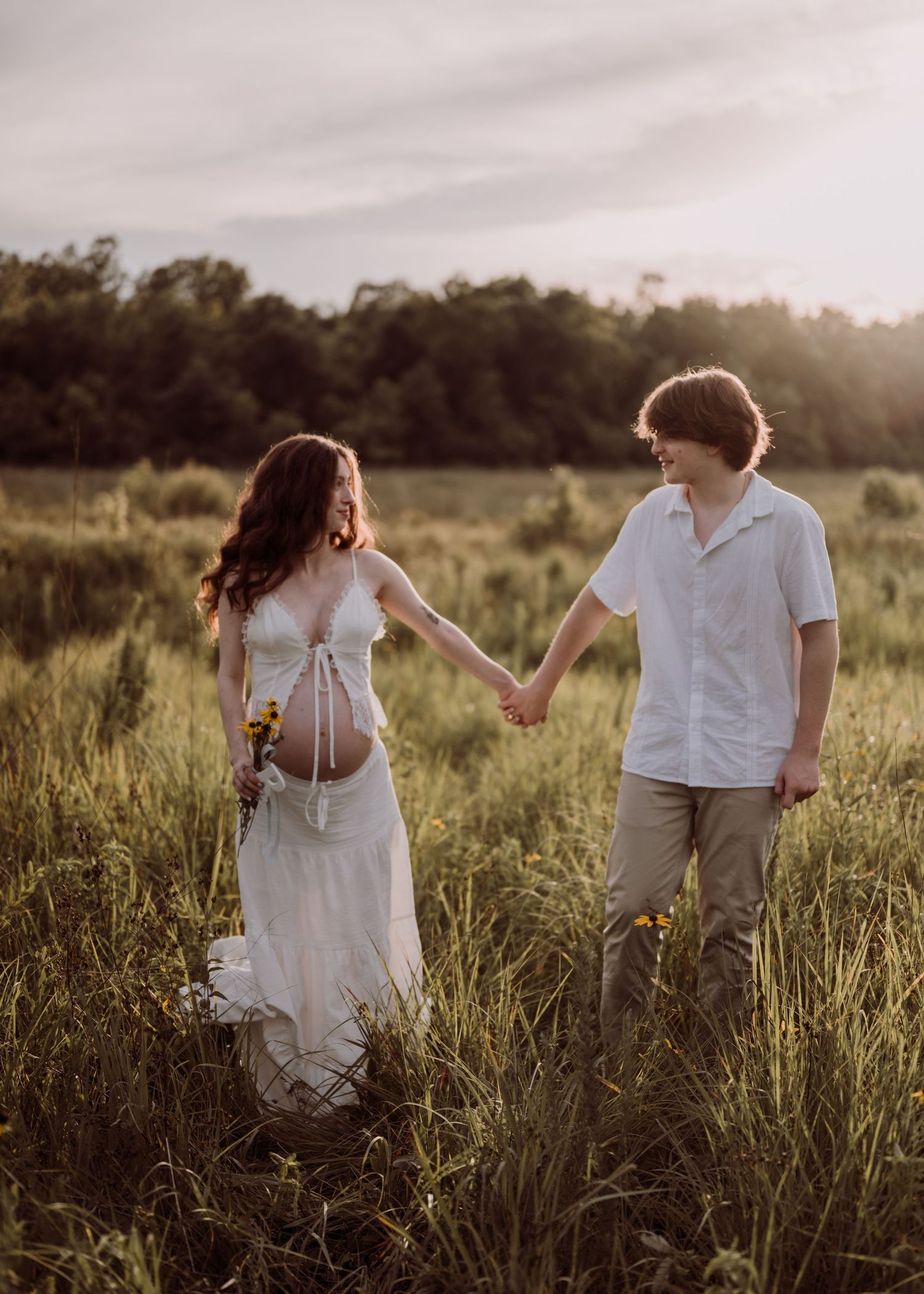 A pregnant couple holds hands while walking through a golden meadow at sunset in a romantic maternity photo session.