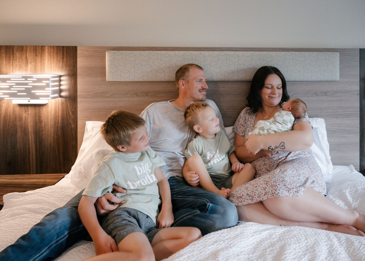 Family of five with newborn baby relaxing together on a hotel bed, smiling and bonding in a warm, cozy setting.