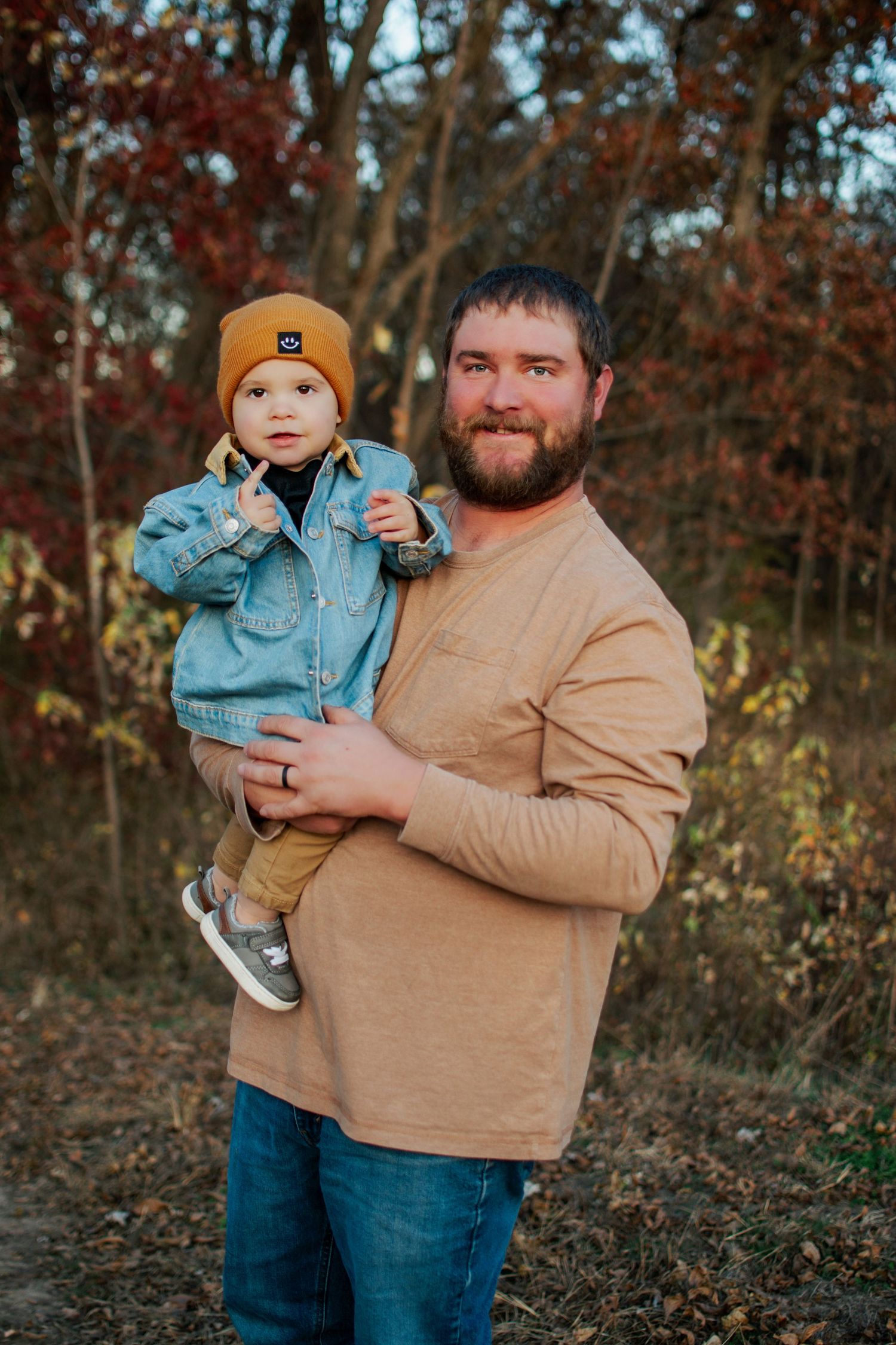 Smiling bearded dad holding toddler in mustard beanie and denim jacket outdoors with colorful fall foliage.