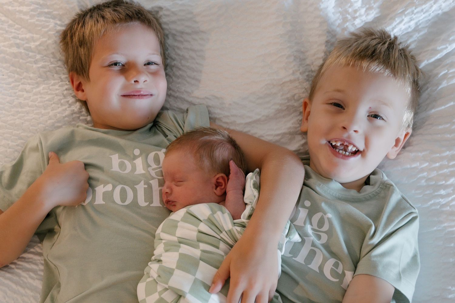 Two big brothers in matching green shirts smile with their newborn sibling lying between them on a white bed.