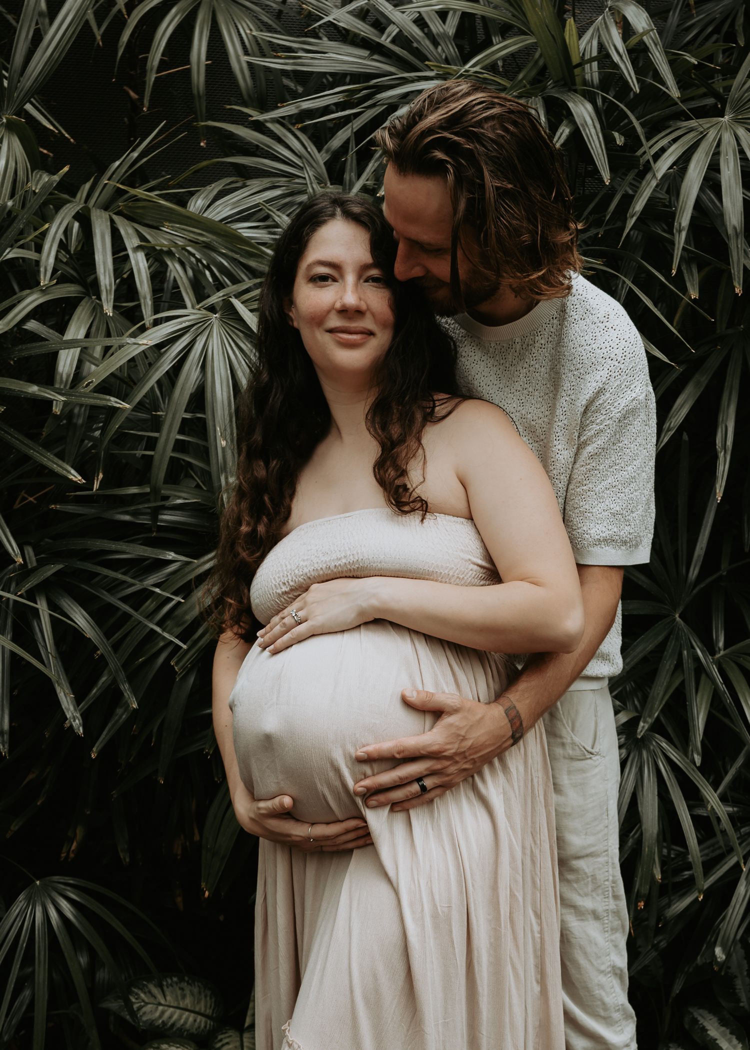 Couple poses for maternity photo shoot in front of lush tropical palms, man embracing pregnant woman in flowing dress.