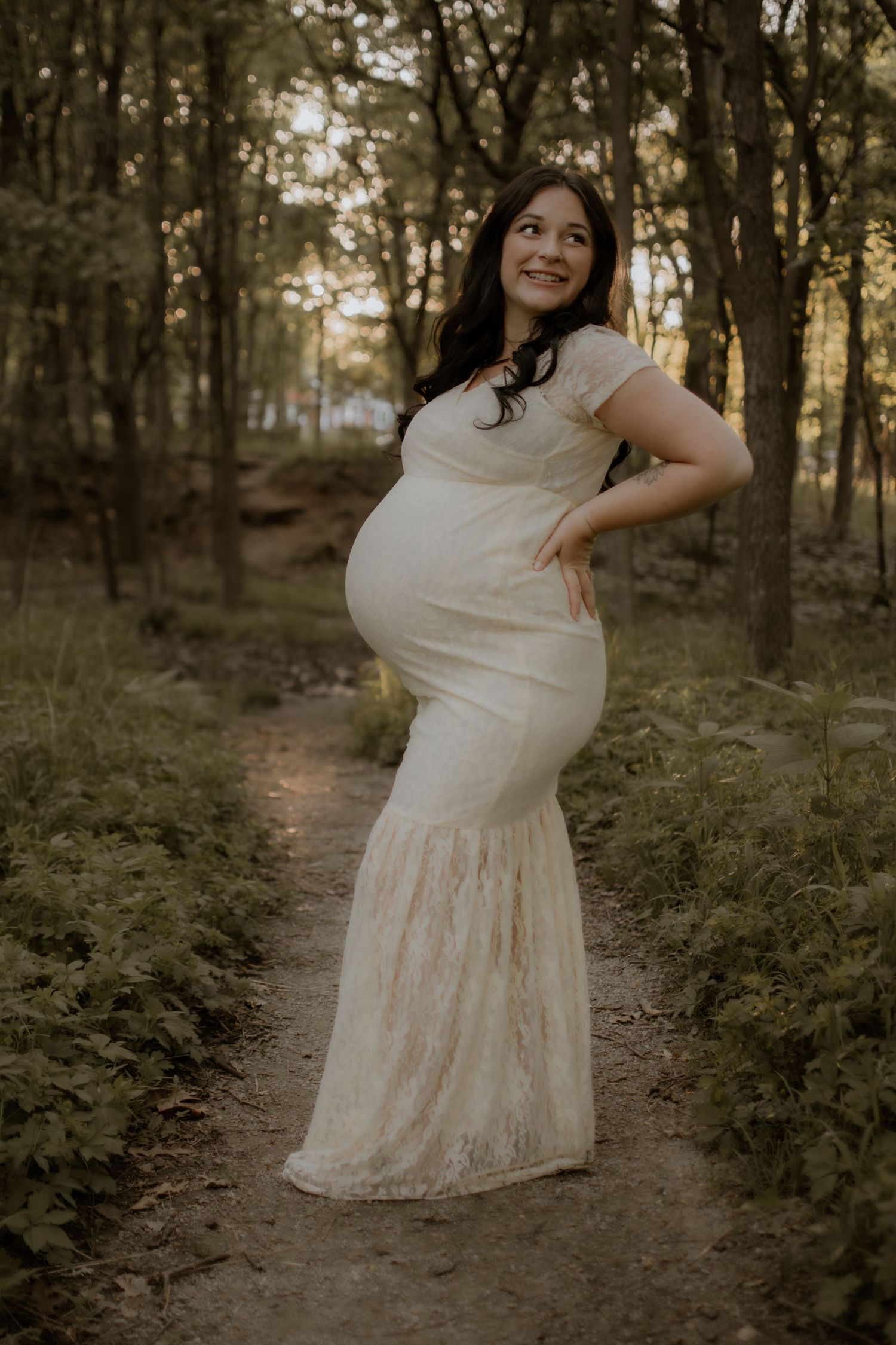 Smiling pregnant woman in cream lace fitted gown posing on a forest path at dusk.