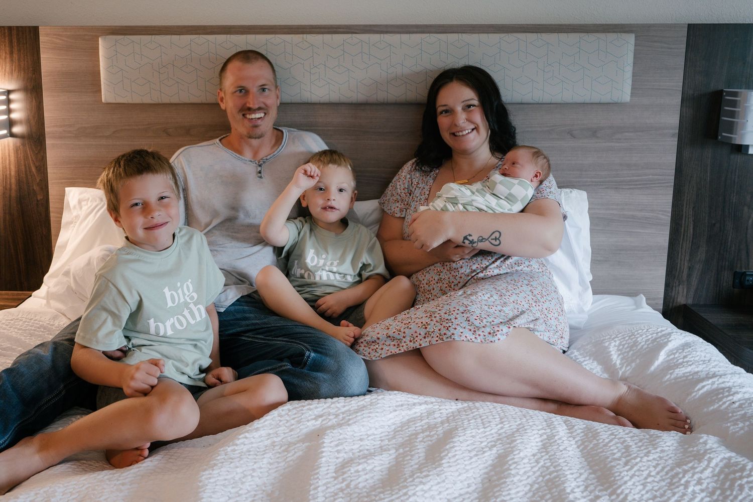 Happy family of five poses on a bed, parents with three young boys and a newborn baby, smiling together.