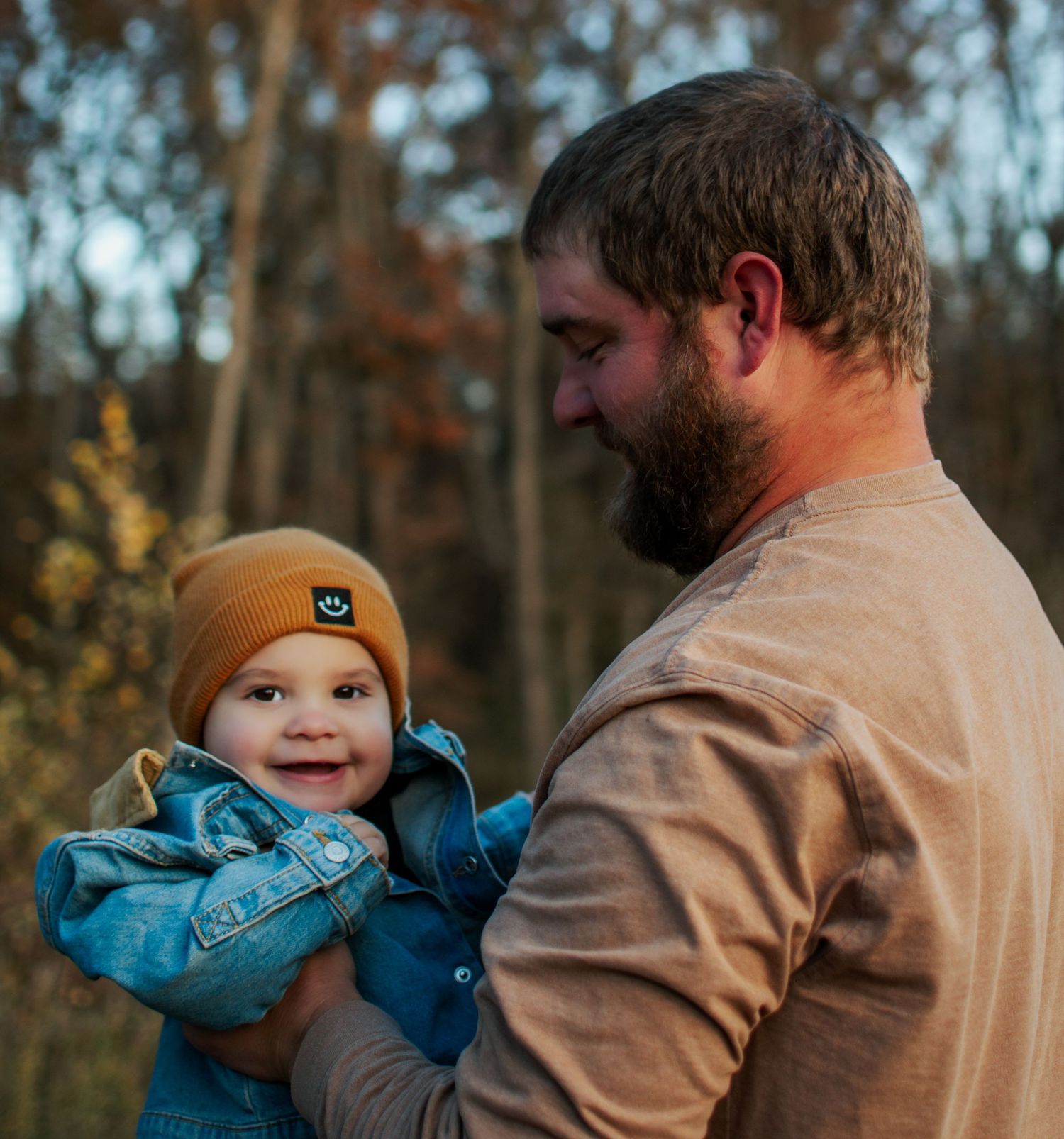 Bearded father smiling at happy baby in mustard beanie and denim jacket in a fall outdoor setting.