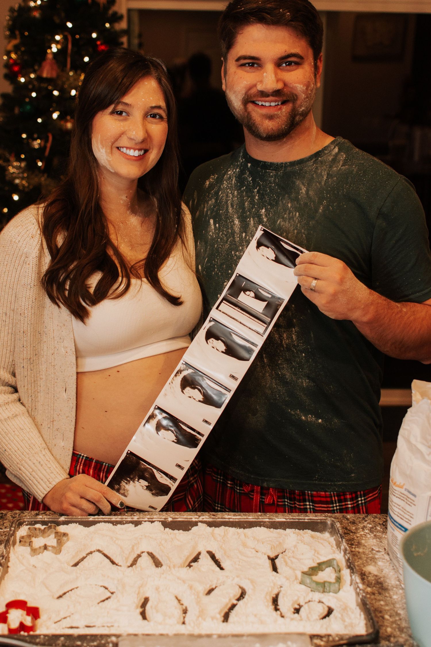 Smiling couple holding ultrasound strip beside flour-covered baking sheet pregnancy announcement.