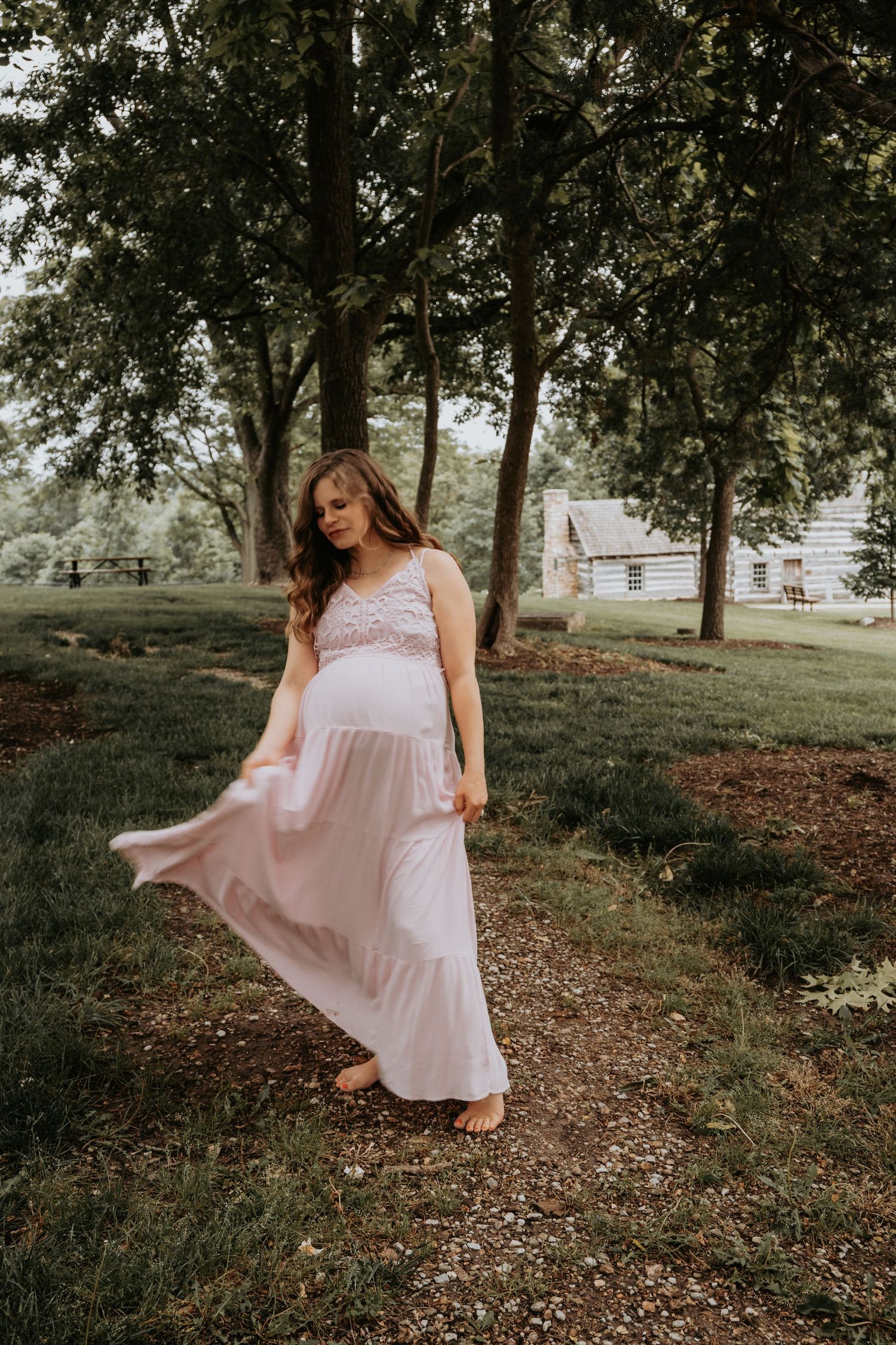 Pregnant woman in blush pink maxi dress walking barefoot on a tree-lined path outdoors.