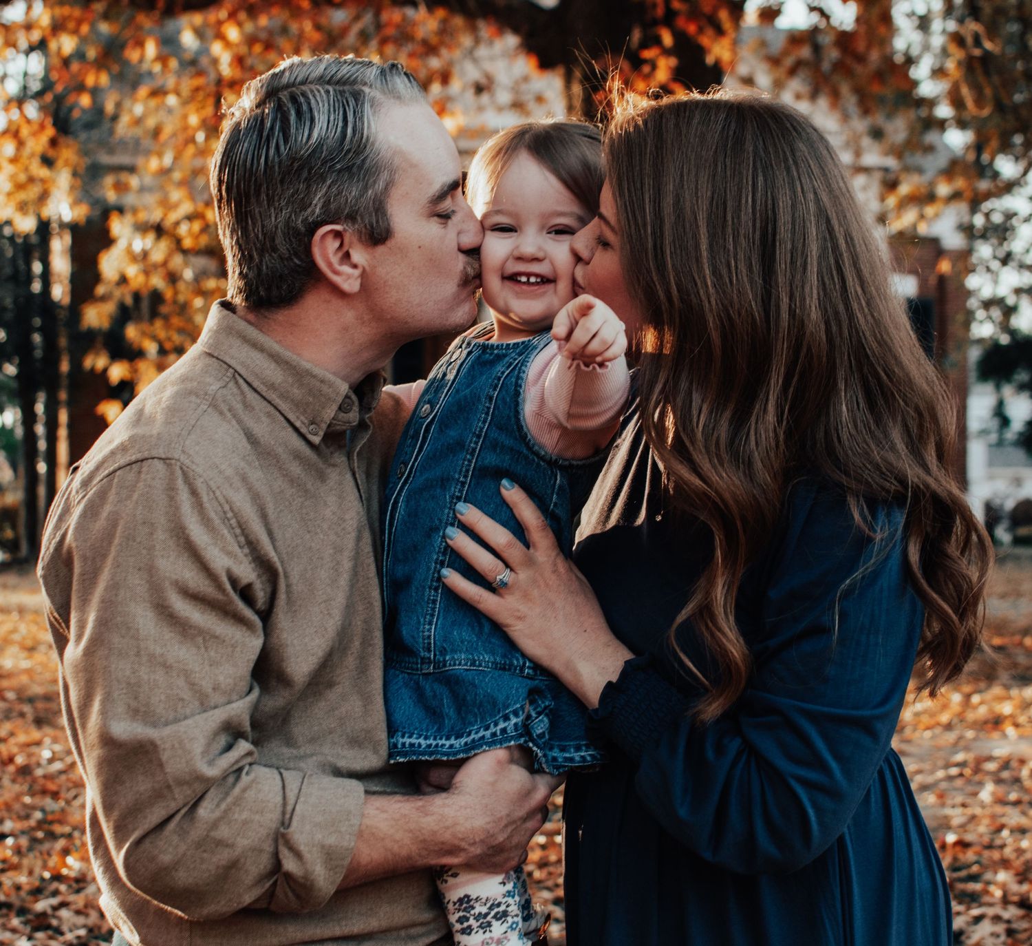 Parents lovingly kiss their giggling baby on both cheeks during a fall family photo session.