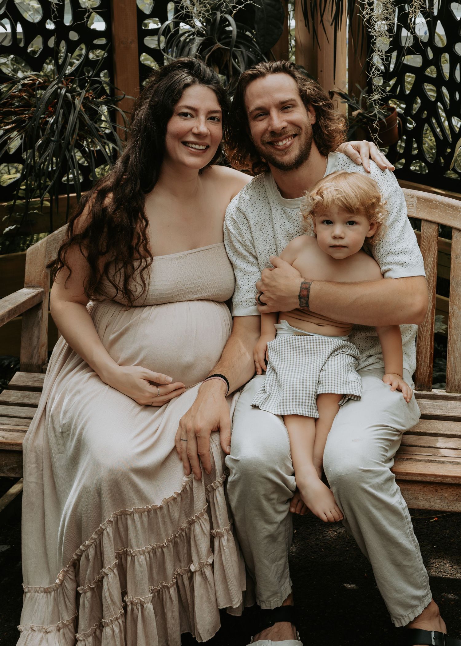 A happy pregnant woman and her partner sit on a bench with their young child in a tropical garden.