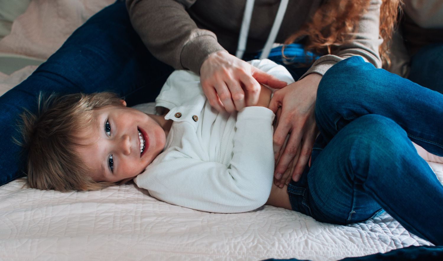 Happy toddler boy in white outfit lying on bed being tickled by parents in cozy home setting.