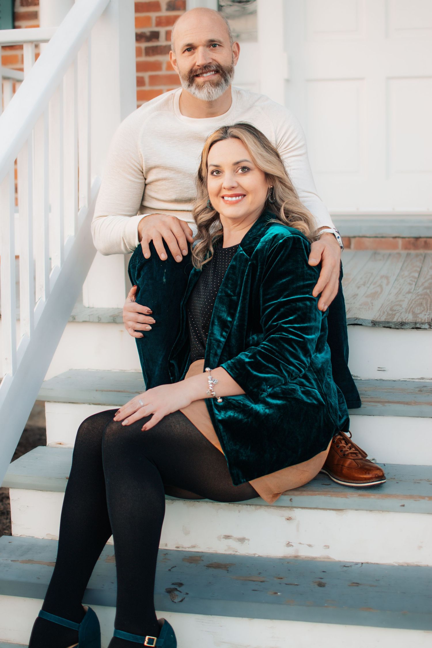 A couple sits closely together on white porch steps, smiling warmly during an outdoor photo session.