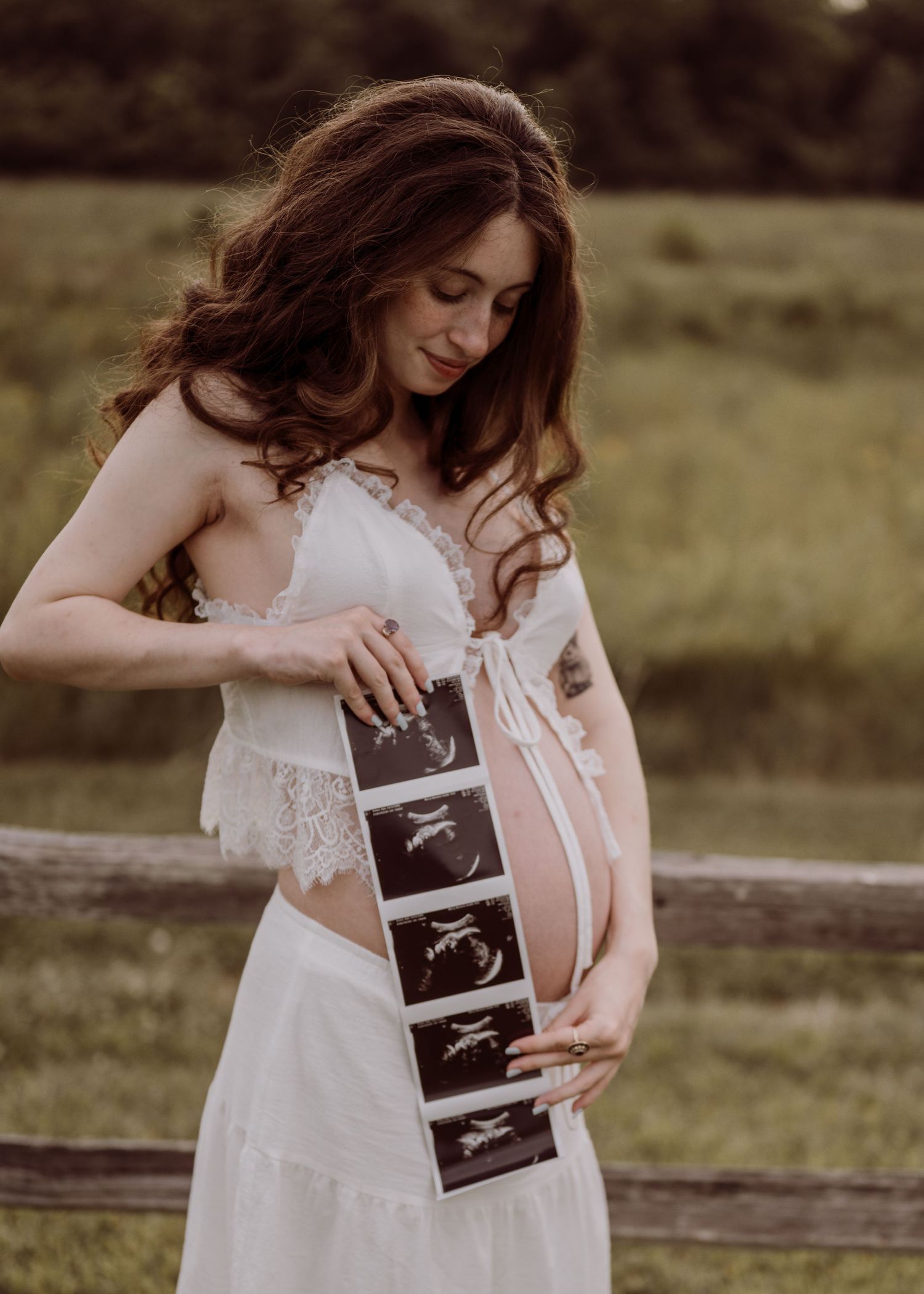 Pregnant woman in white lace dress holding ultrasound photos against her baby bump outdoors near a wooden fence.
