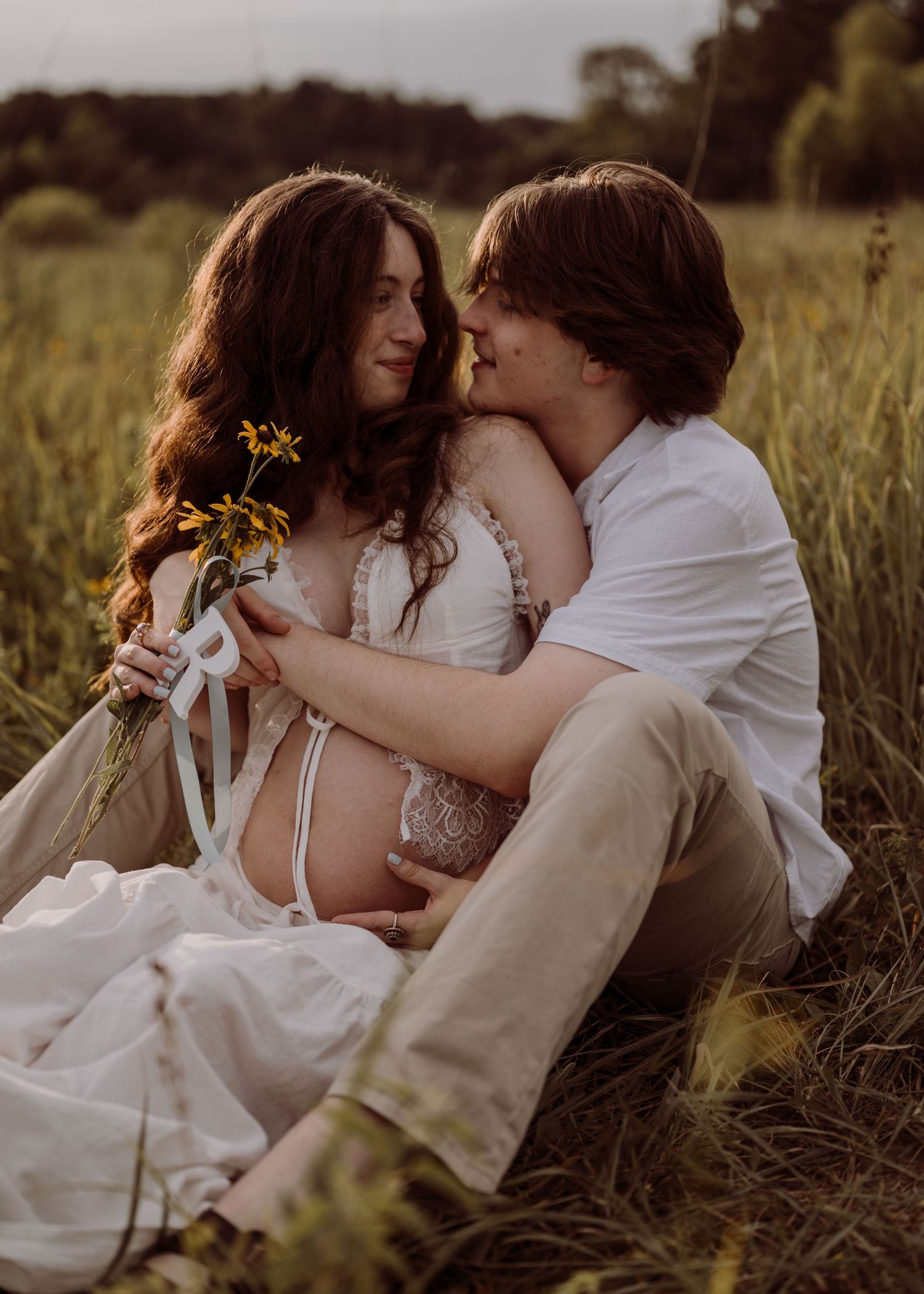 A couple sits intimately in a golden field, the man embracing his pregnant partner holding wildflowers at sunset.