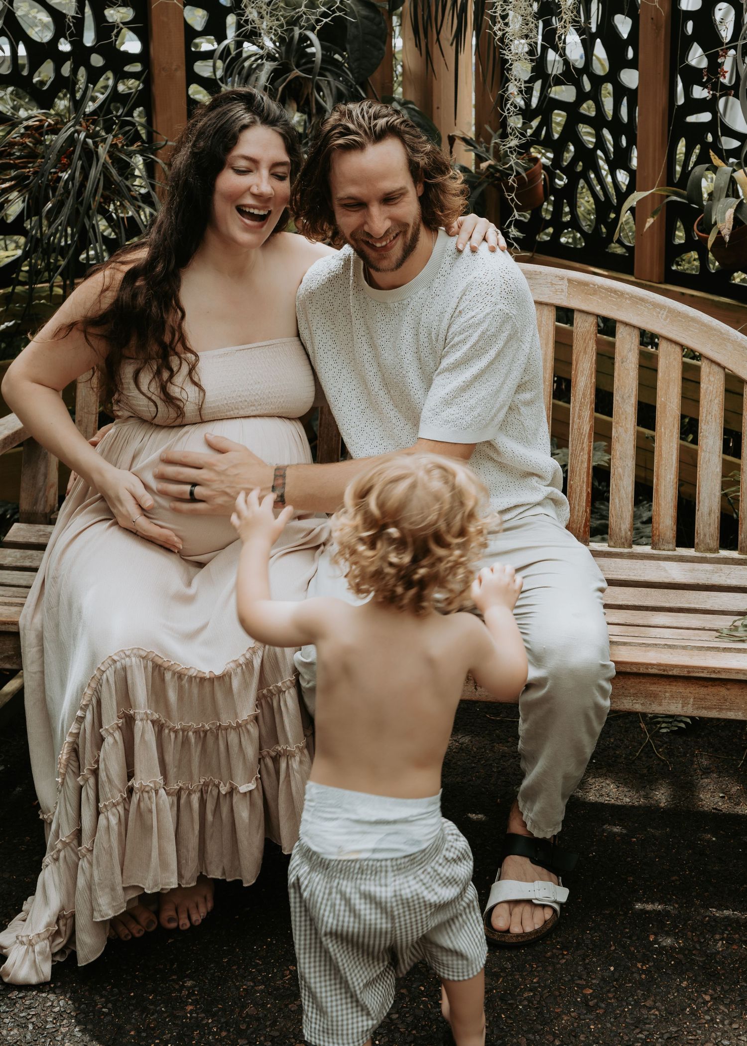 Laughing pregnant woman with partner and toddler on garden bench during family maternity session.