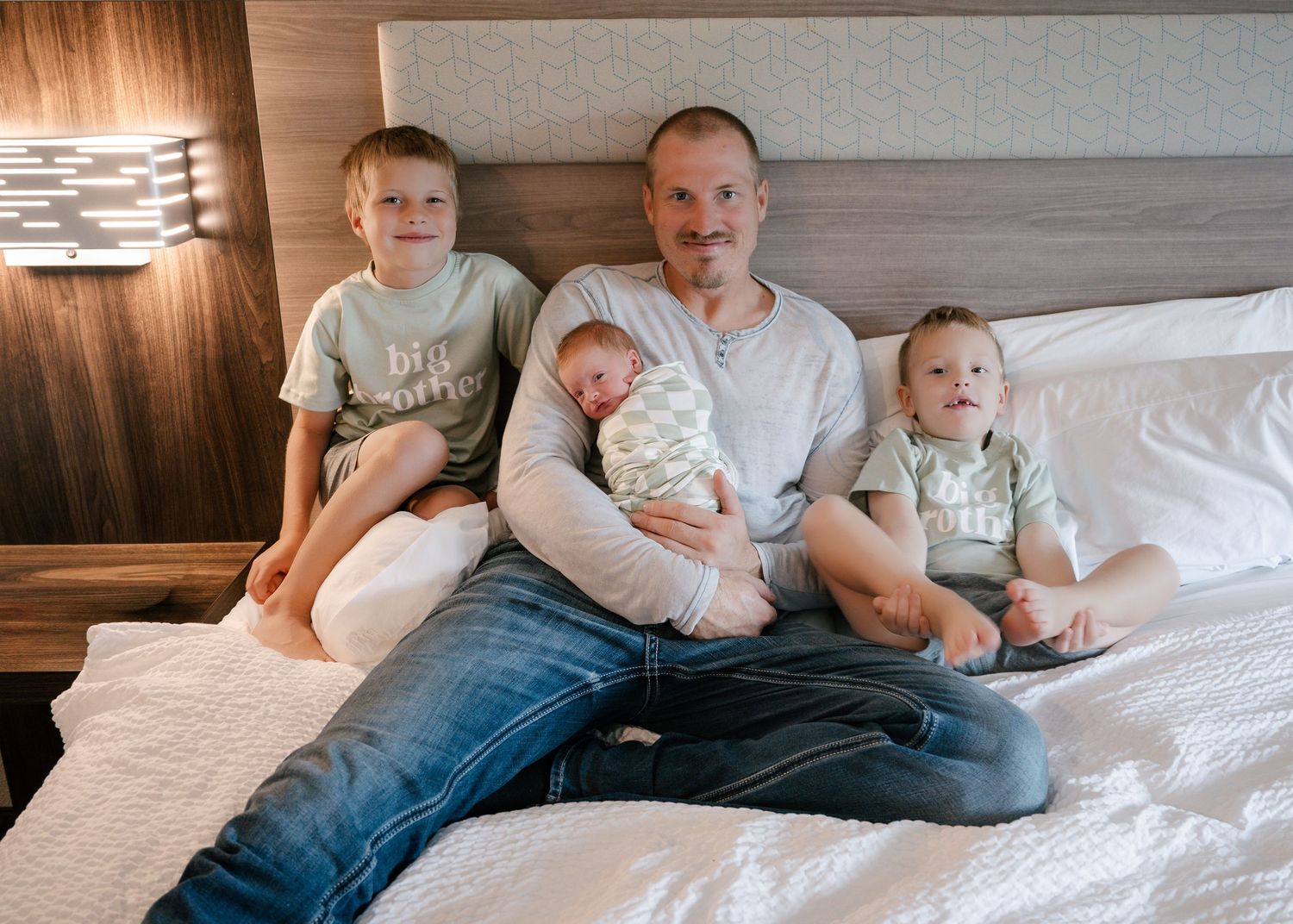 Man sitting on bed holding newborn baby flanked by two young boys in a cozy hotel room setting.