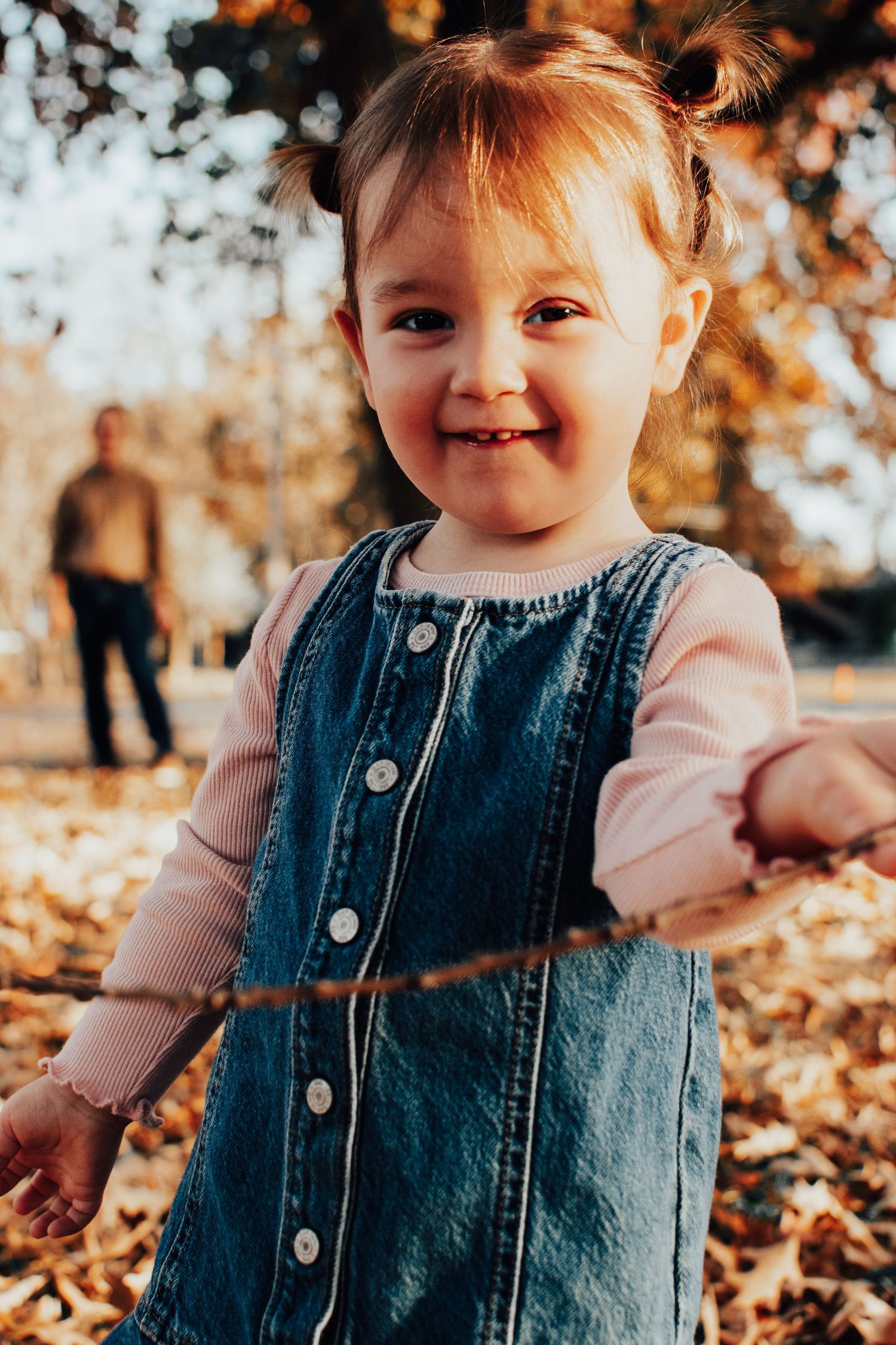 Cheerful toddler girl in denim jumper playing in fallen autumn leaves at golden hour.