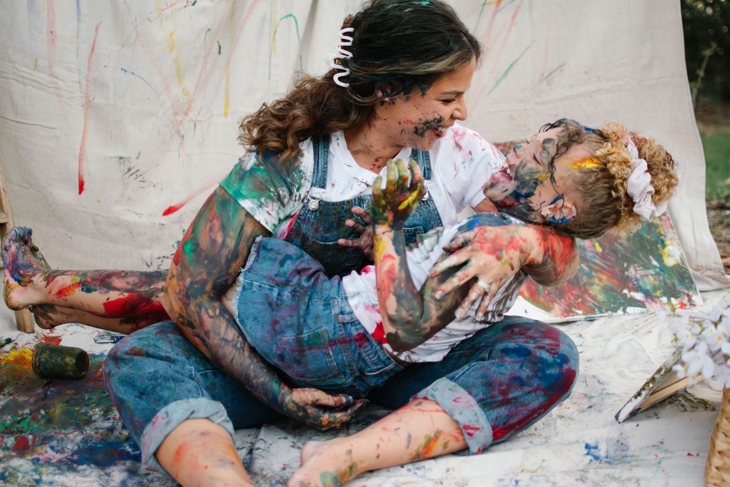 Two paint-covered people laughing and embracing during a fun, colorful paint play session outdoors.
