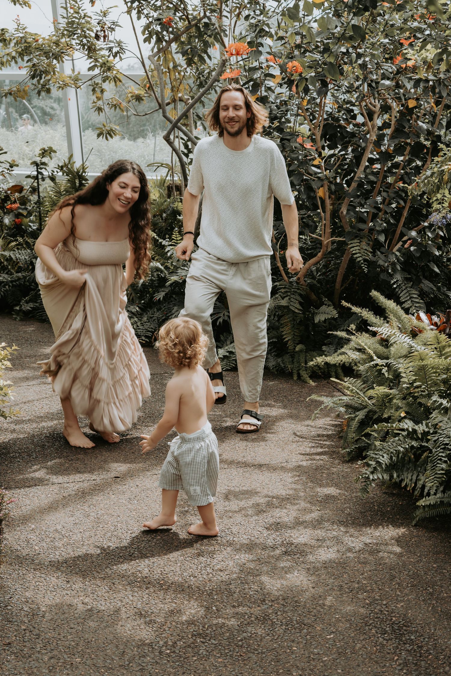A young family with a toddler walks through a lush greenhouse garden, smiling and enjoying nature together.