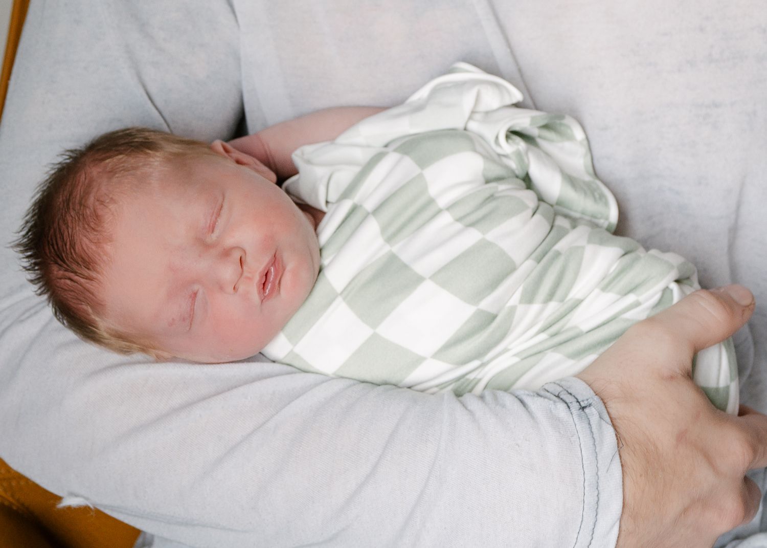 Newborn baby sleeping peacefully, swaddled in a green checkered blanket, cradled in an adult's arms.