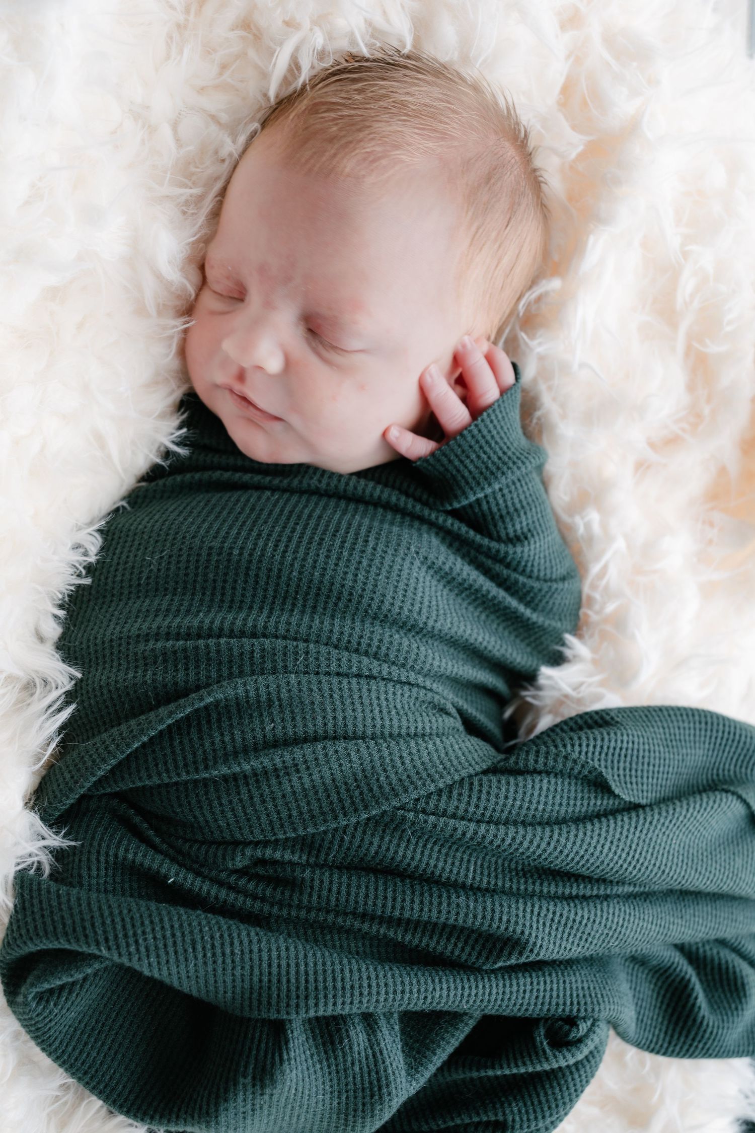 Sleeping newborn baby swaddled in a dark green knit wrap, resting peacefully on a soft white fluffy blanket.