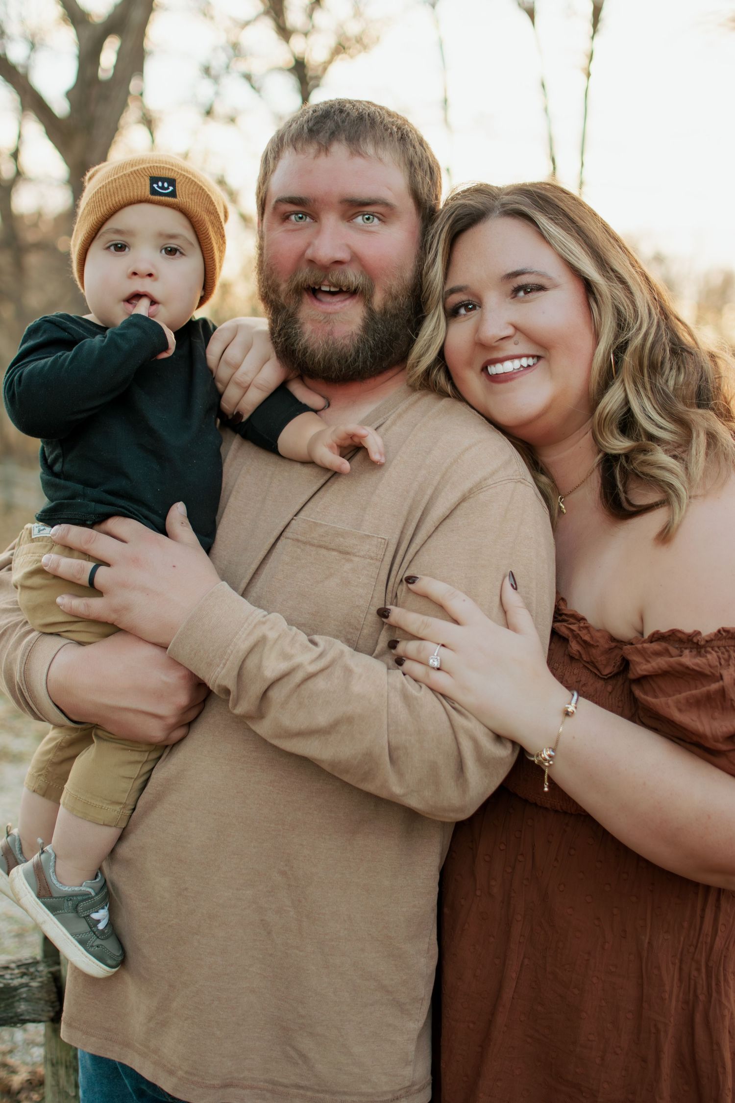 Happy family portrait with bearded dad holding toddler in mustard beanie, mom in rust dress, outdoors in autumn.