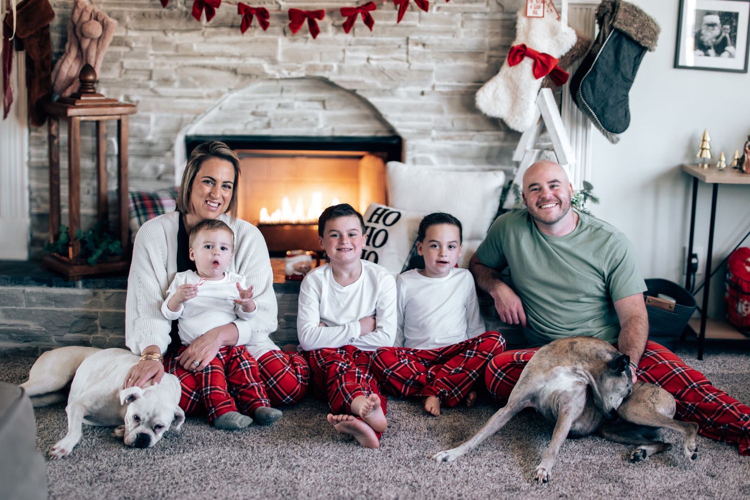 A festive family in matching Christmas pajamas poses by a fireplace with two dogs.