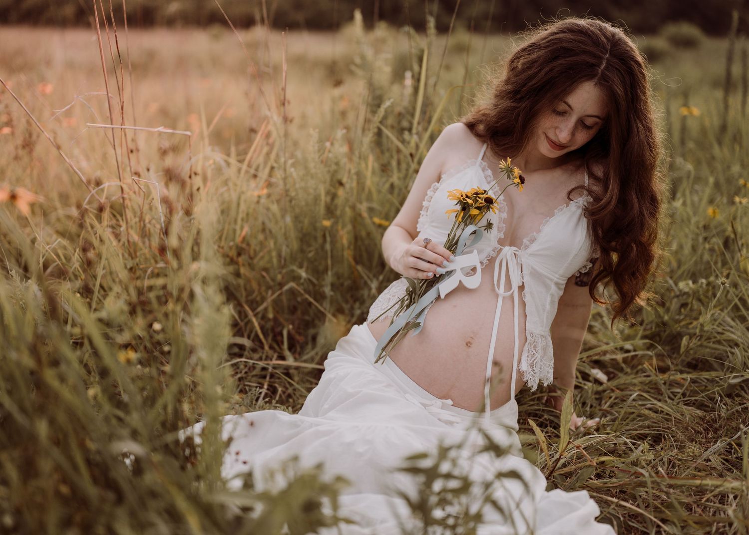 Pregnant woman in white dress sitting in golden field holding yellow wildflowers during sunset maternity photoshoot.