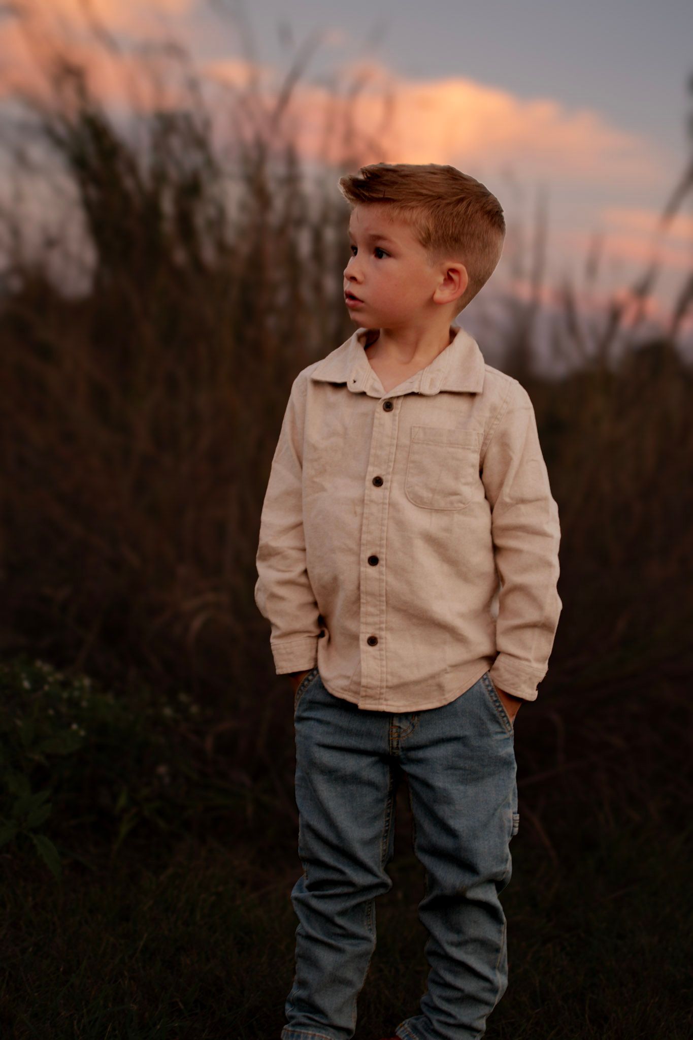 Young boy in beige linen shirt and jeans standing outdoors at sunset with dramatic pink sky behind him.