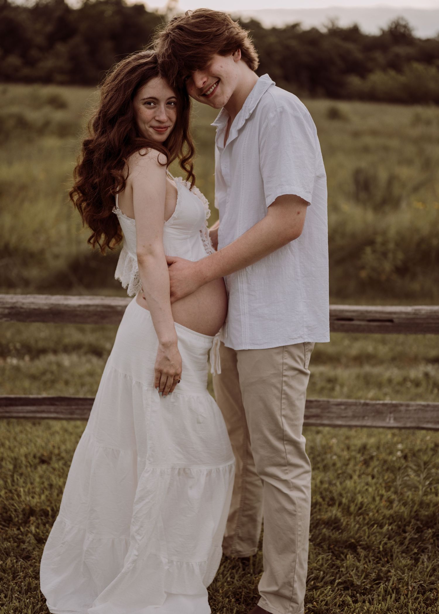 A smiling pregnant woman in a white dress poses with her partner in a lush green outdoor maternity portrait.