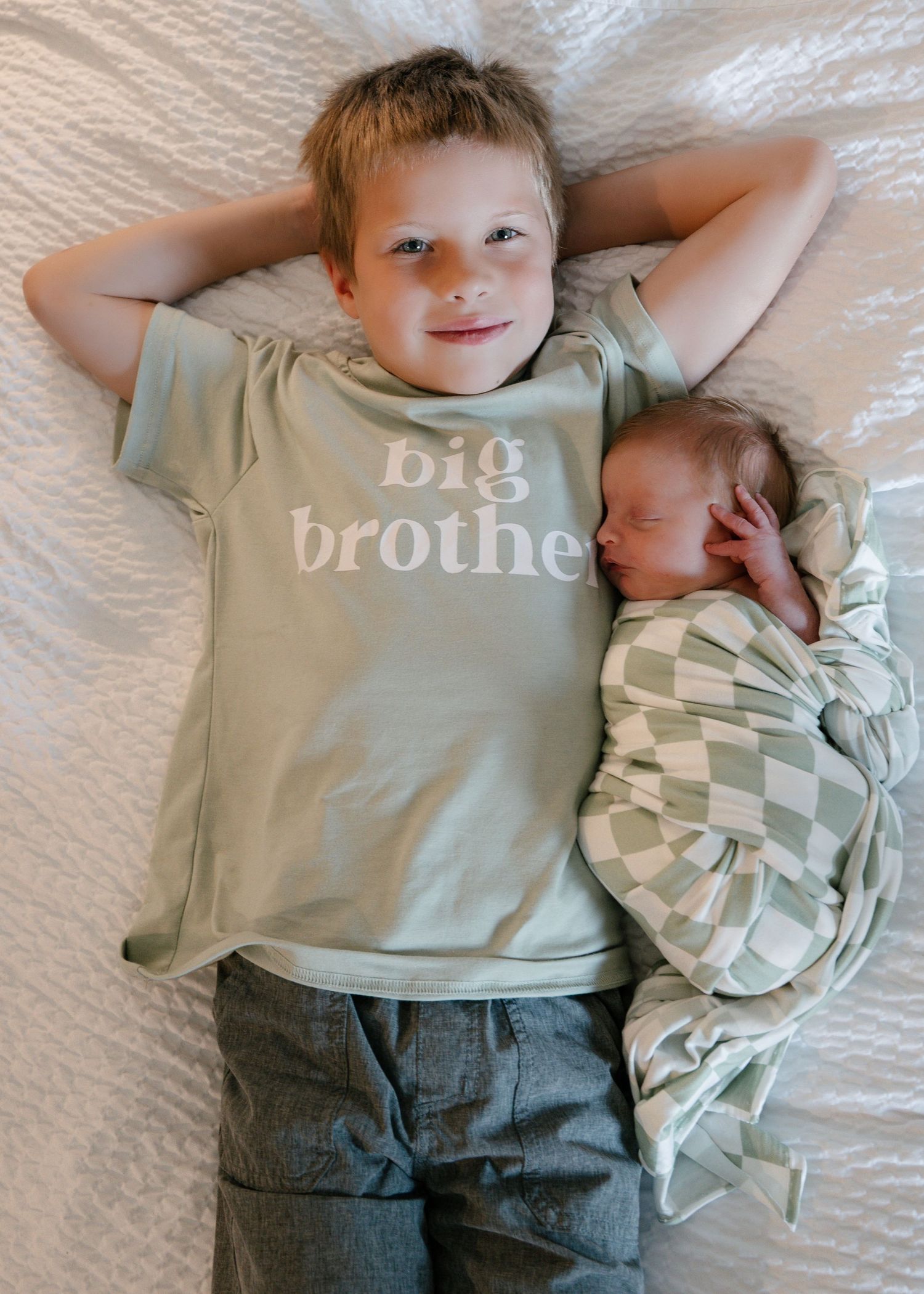 A young boy wearing a sage green big brother shirt lies next to a sleeping newborn wrapped in a checkered blanket.