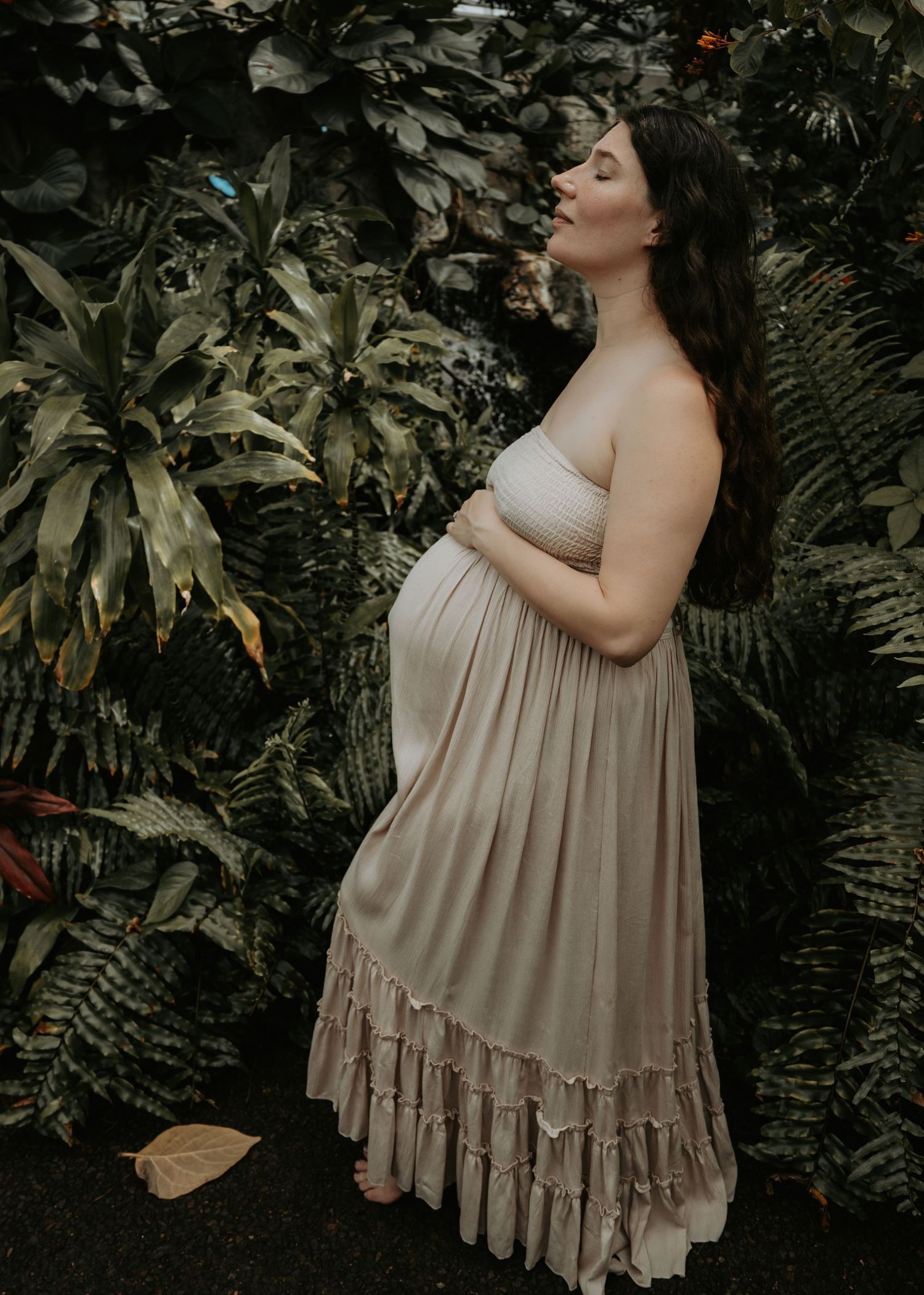 Pregnant woman in a flowing beige ruffled maxi dress standing among lush tropical greenery, cradling her baby bump.